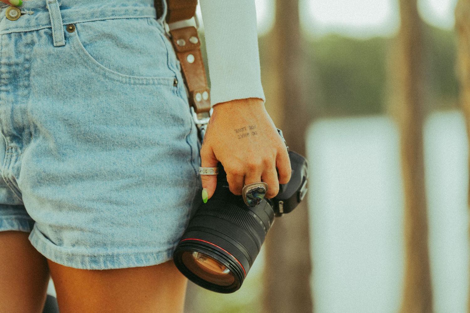 Woman in denim shorts holding DSLR camera with hand tattoo visible.
