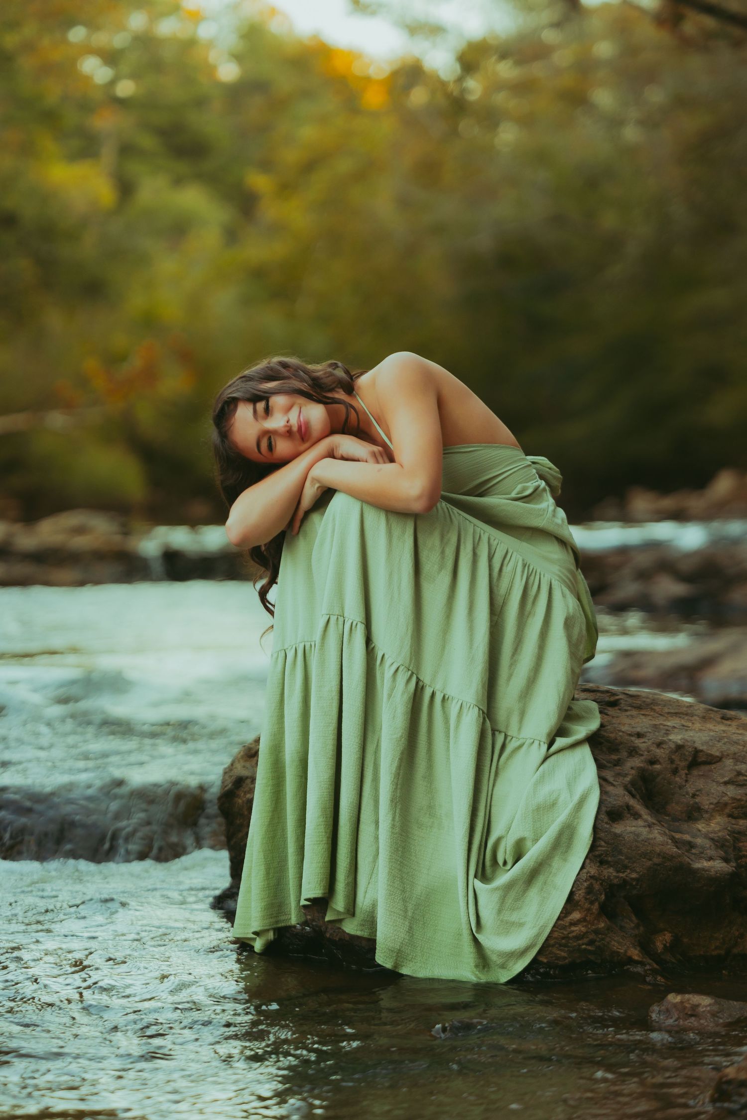 Woman in sage green dress poses dreamily on rocks by a flowing river surrounded by autumn foliage.