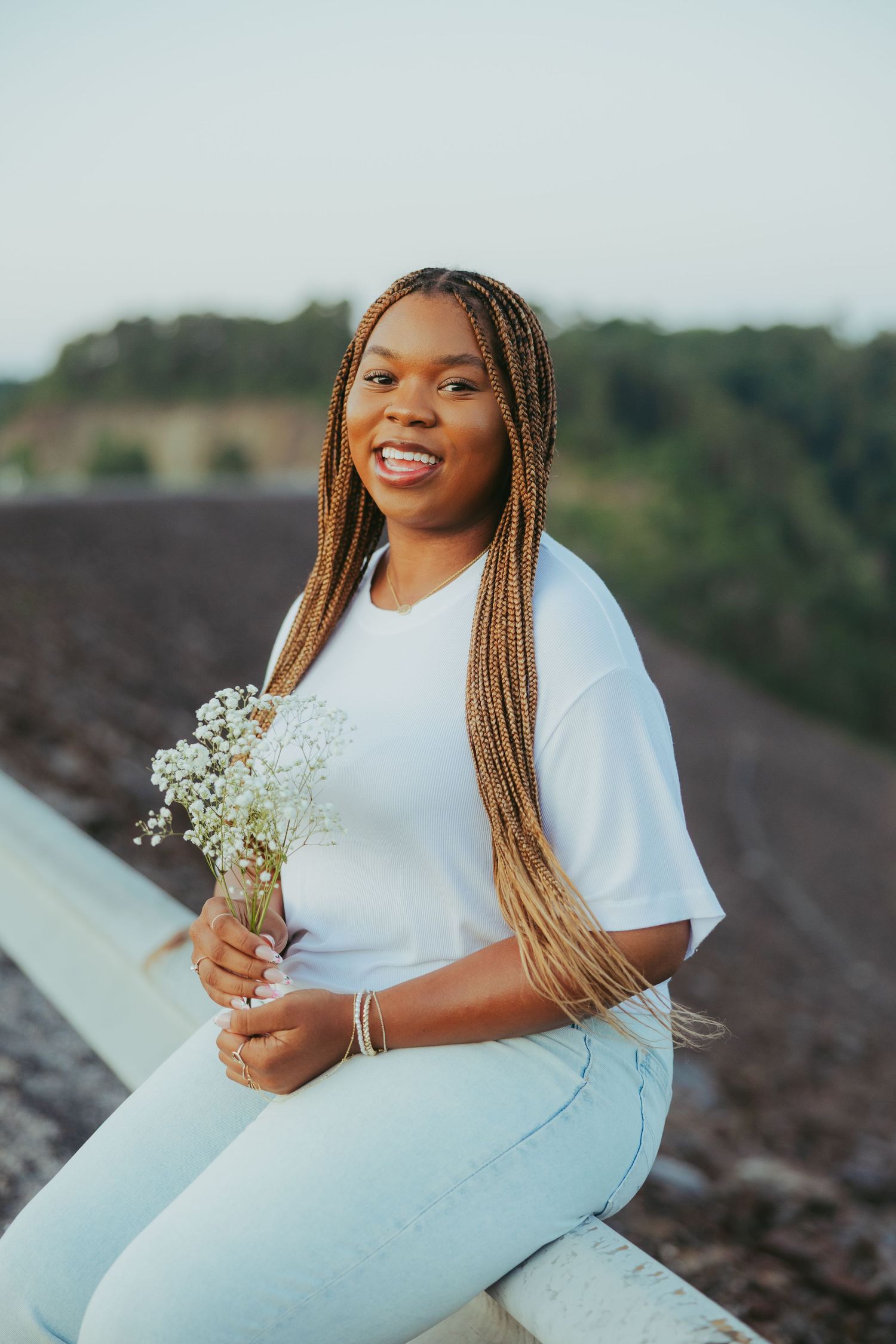 Smiling young Black woman with long braids holding white flowers sitting outdoors at sunset.