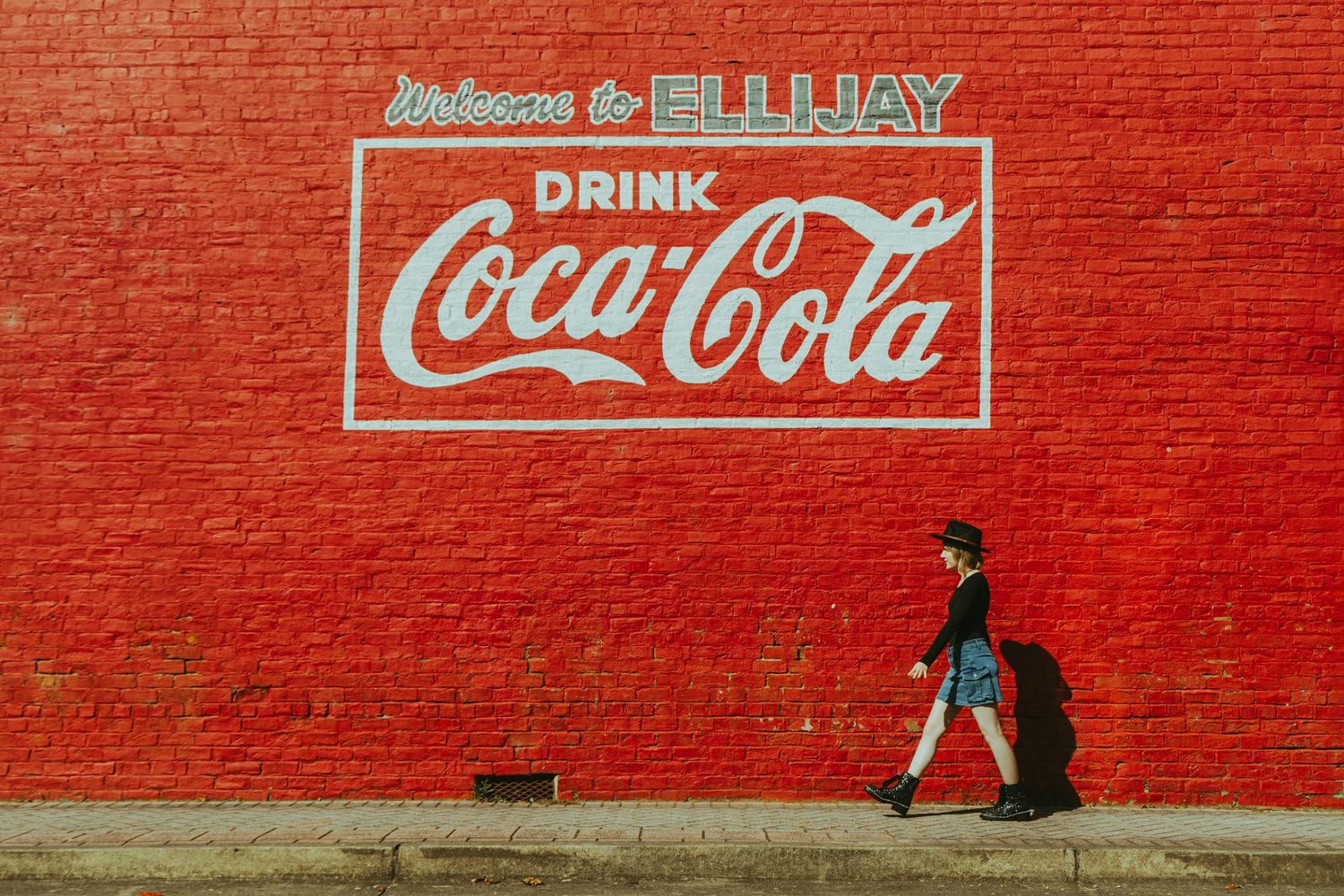 Person walks past vintage red brick wall with classic Coca-Cola mural in Ellijay Georgia.
