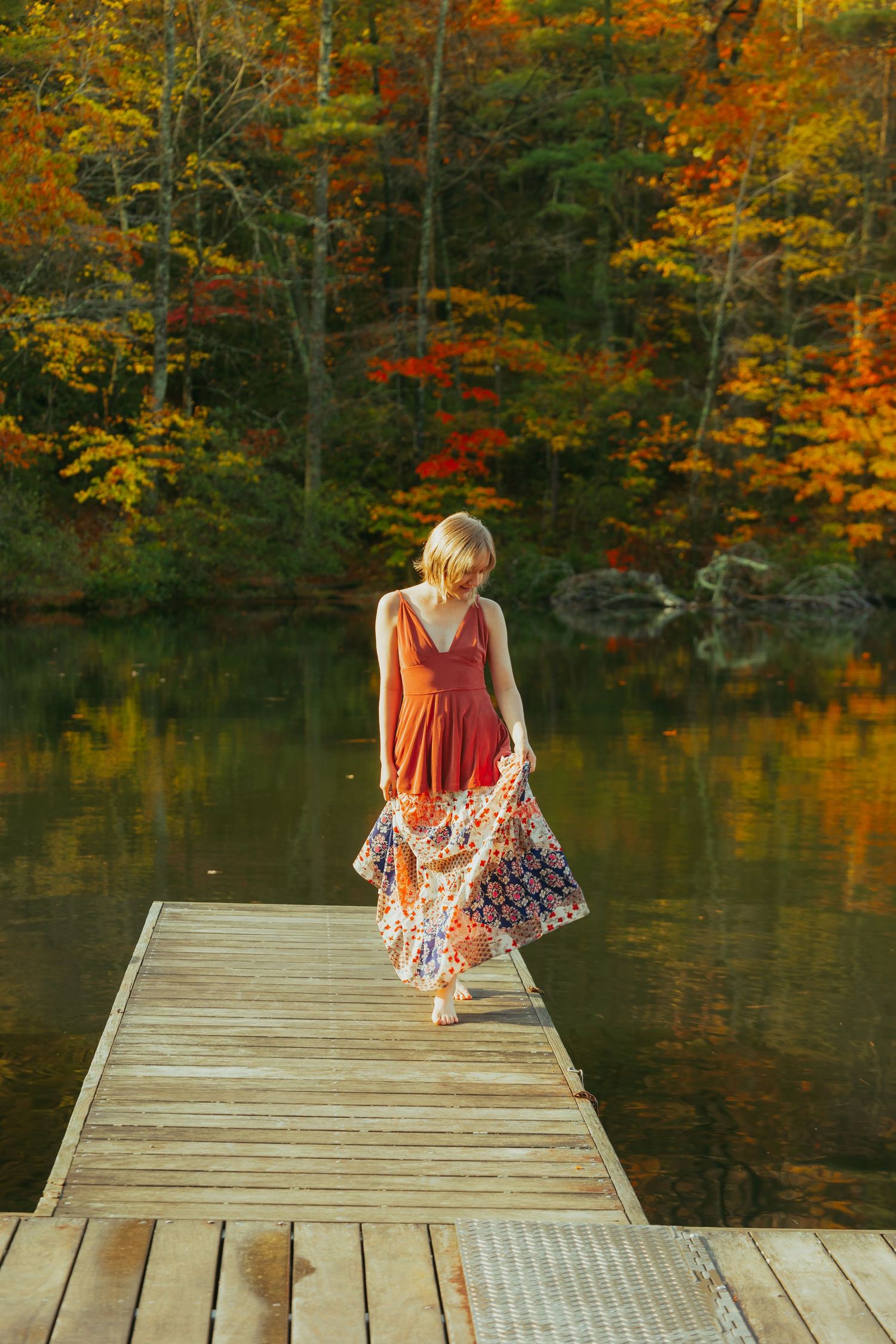 Woman in flowing boho dress walks on wooden dock beside colorful autumn lake reflection.
