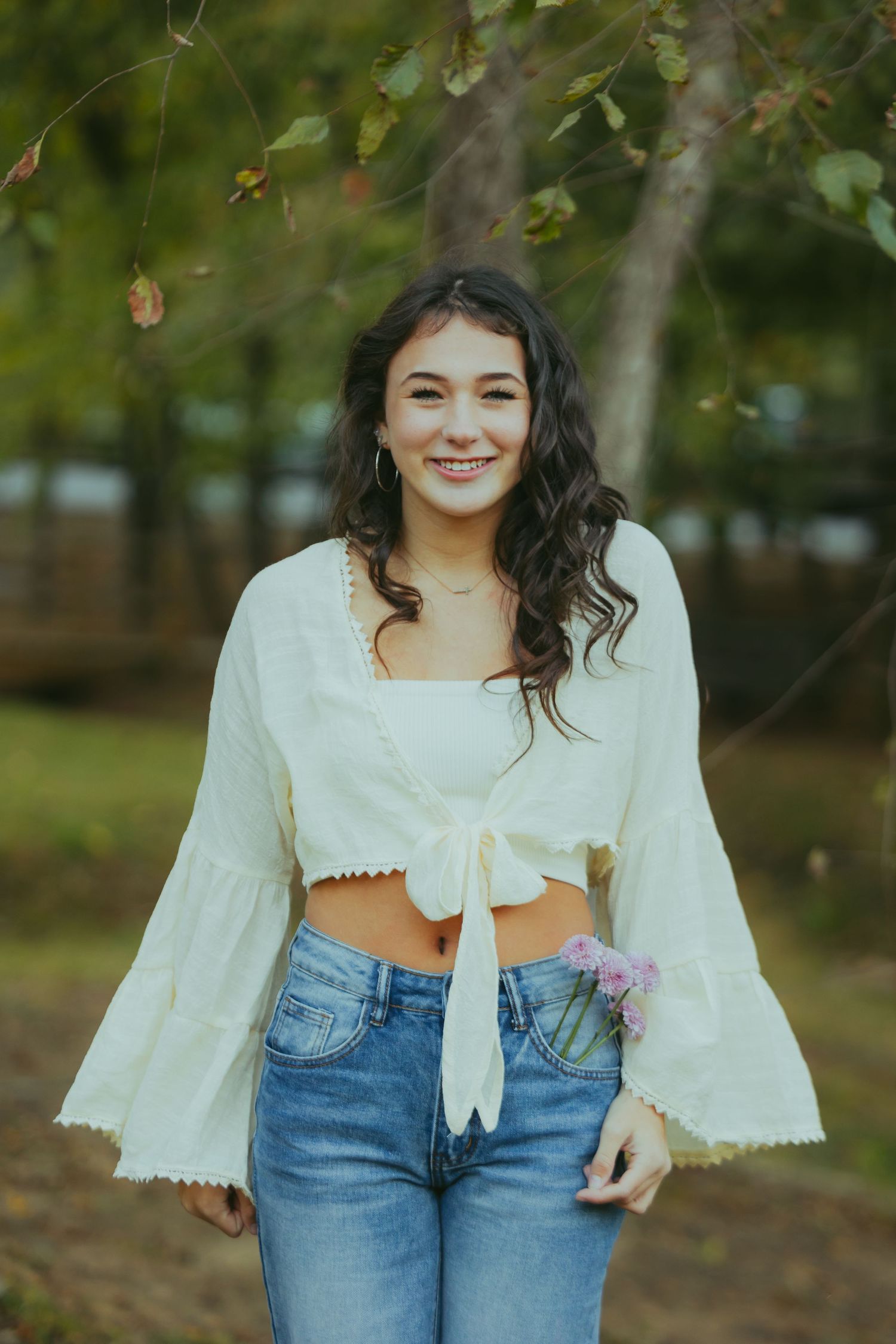 Smiling brunette teen in white crop top and jeans standing outdoors among autumn trees.