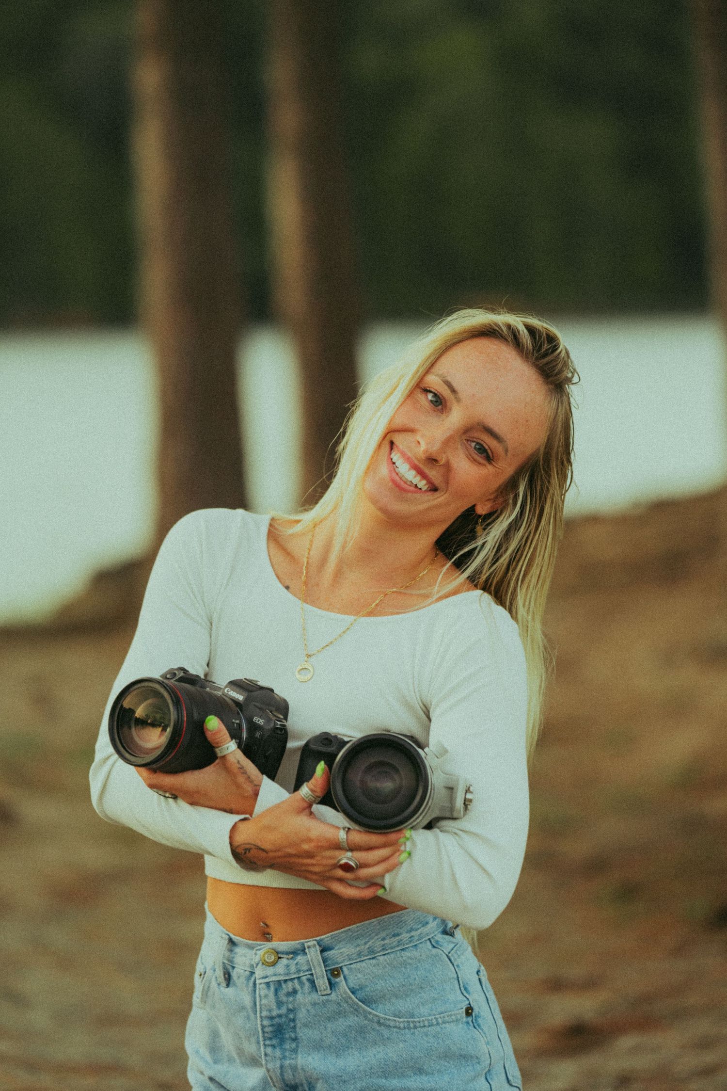 Smiling blonde female photographer holding two DSLR cameras in pine forest.