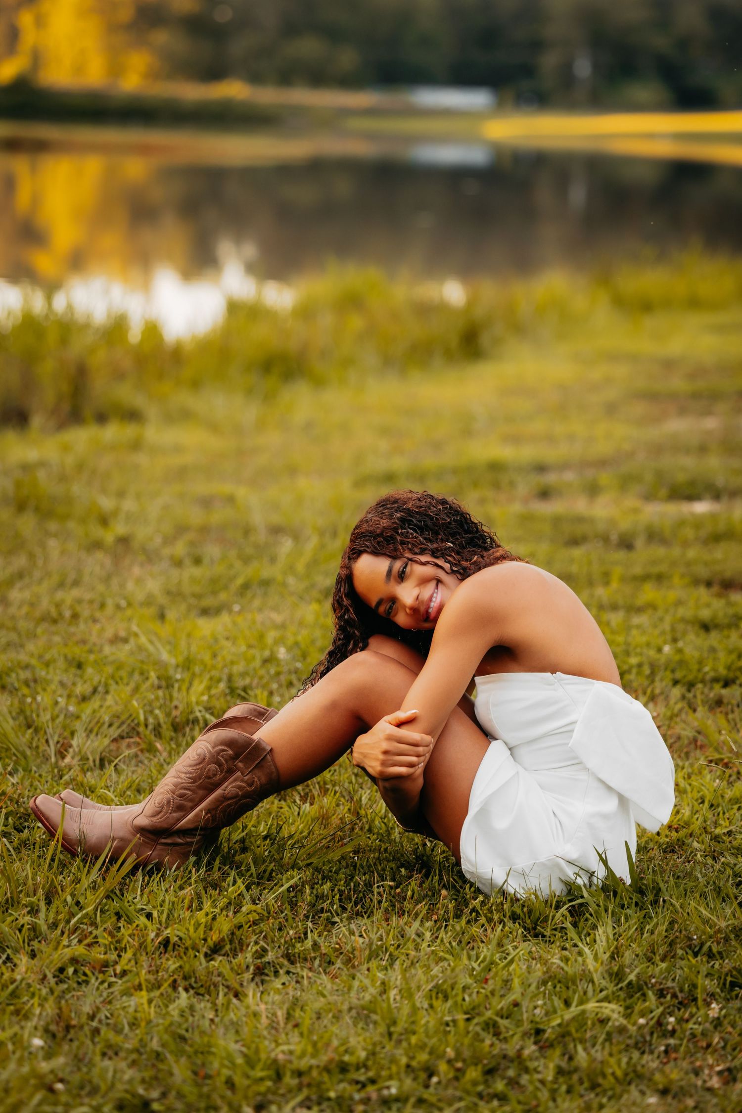 Smiling woman in white dress and cowboy boots sits on grass near a golden autumn pond.