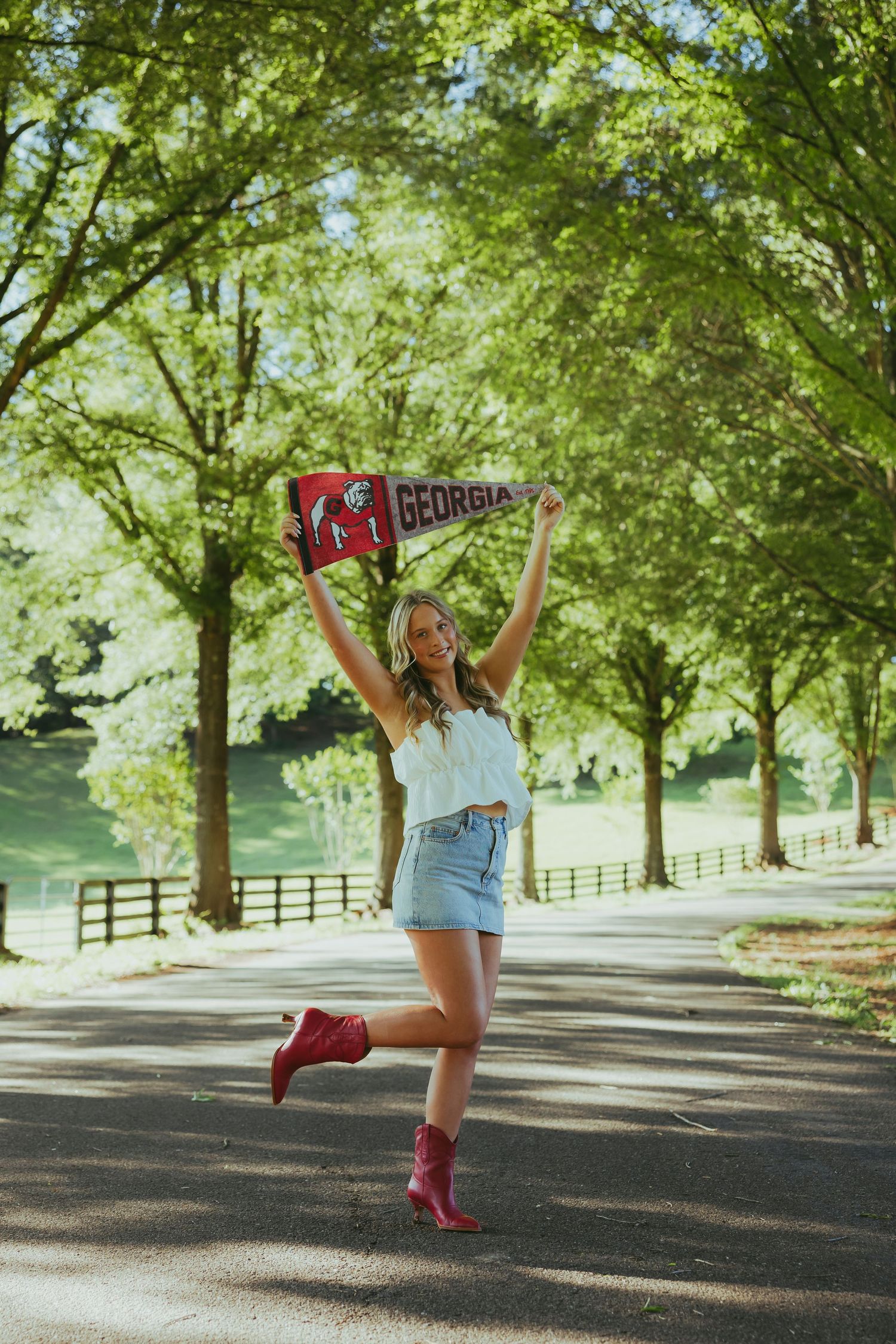 Joyful blonde woman holding Georgia pennant on tree-lined road wearing denim skirt and red boots.