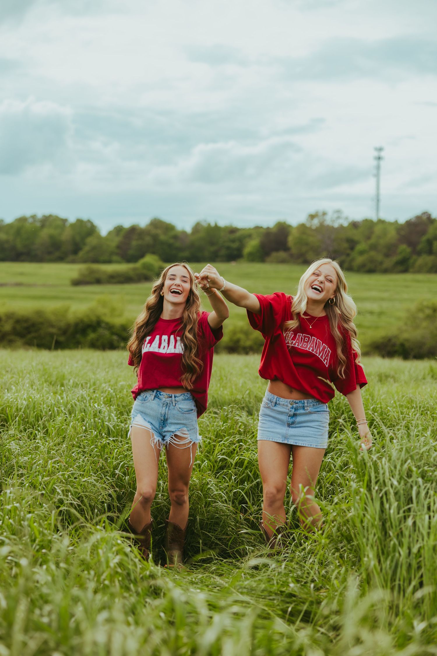 Two laughing girls in red Alabama shirts and denim shorts holding hands in field.