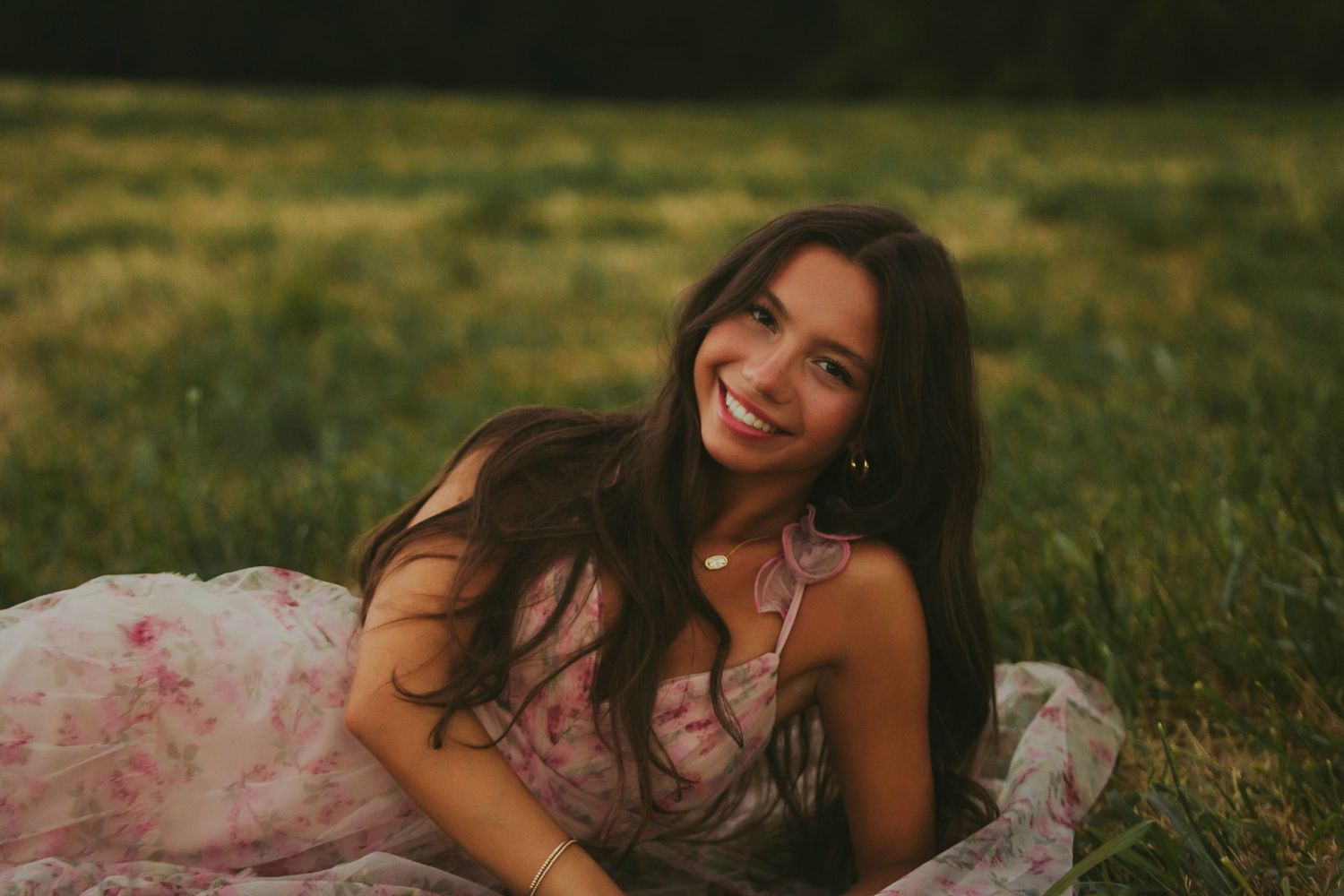Smiling brunette woman in a pink floral dress lying in a green field at golden hour.