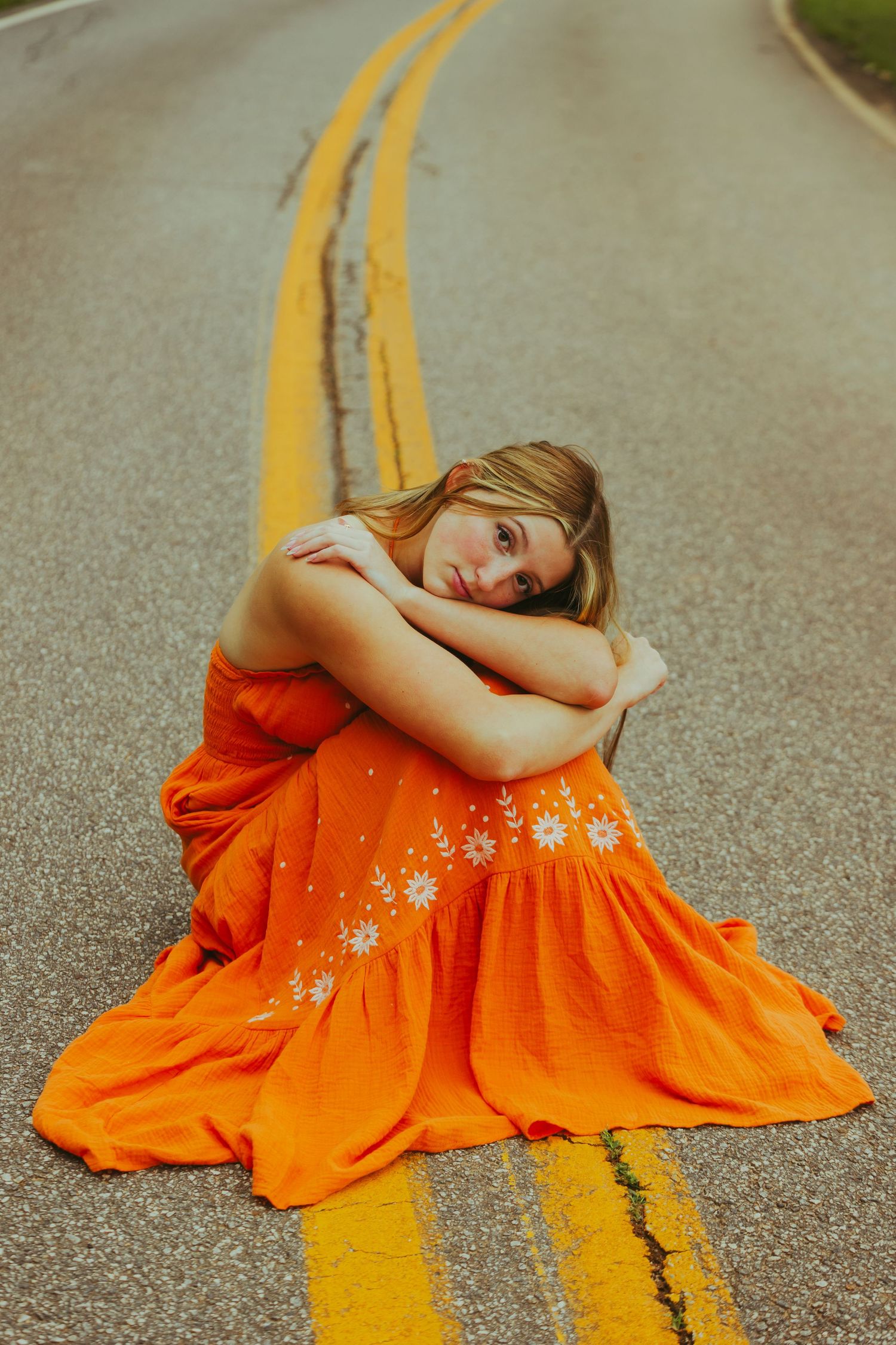 Young woman in orange floral dress sits on road centerline between yellow painted stripes.