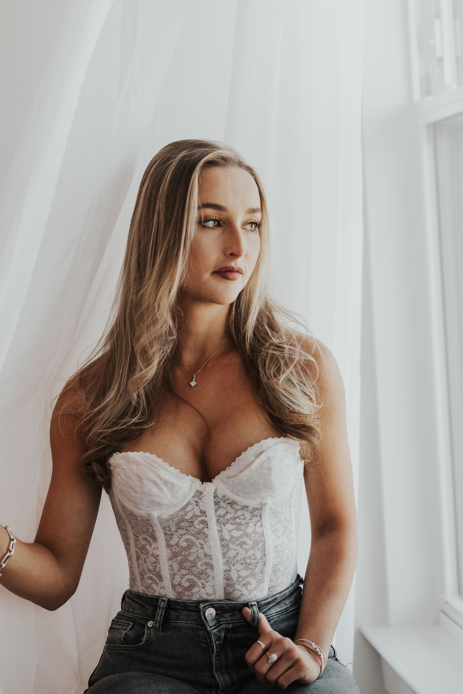 Blonde woman in white lace corset top posing near bright window.