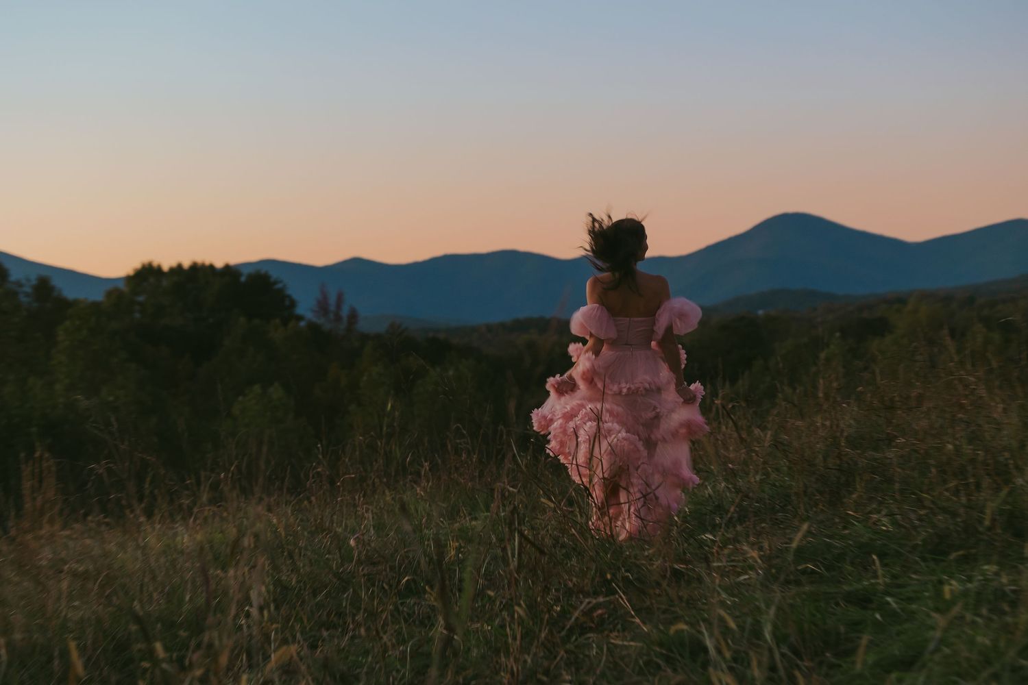 Young woman in pink ruffled gown running through field with mountain sunset backdrop.