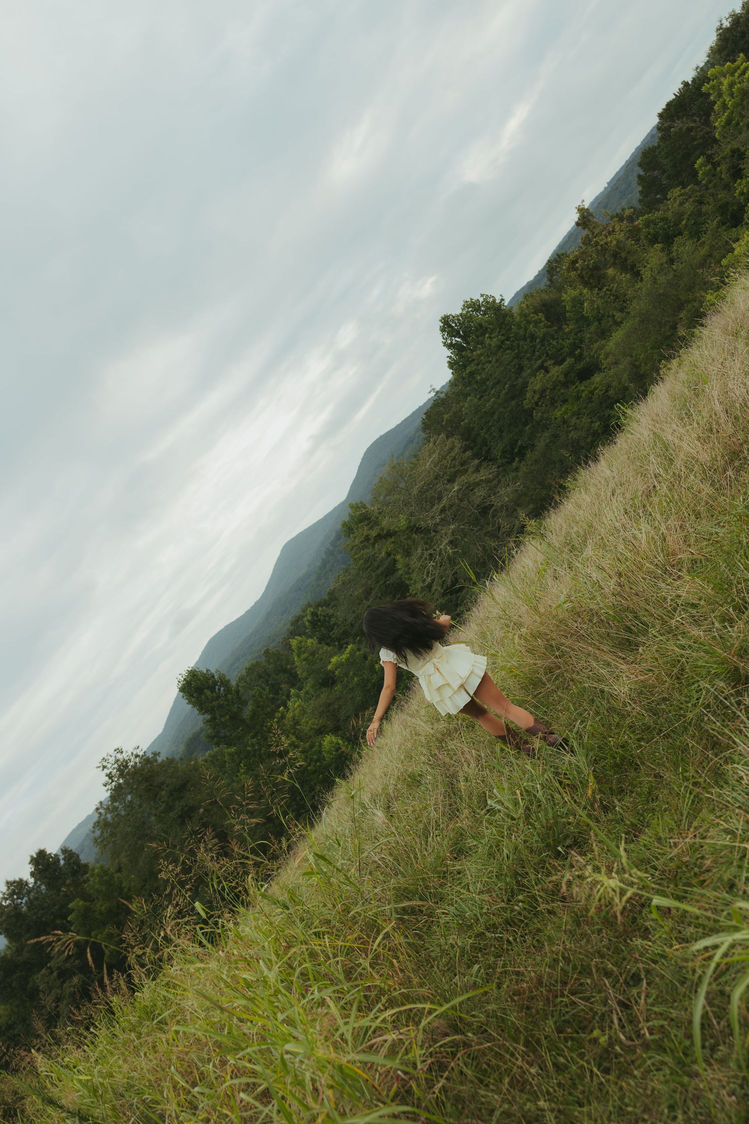 Young woman in white dress climbing steep grassy hillside with mountain landscape background.