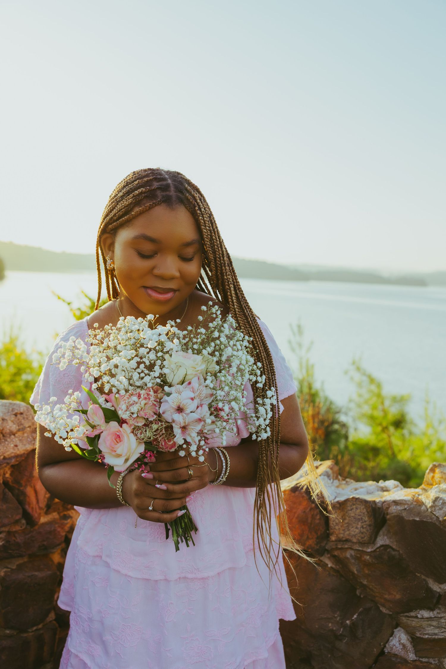Young Black woman with braids holding pink and white flower bouquet outdoors by sunlit lake.