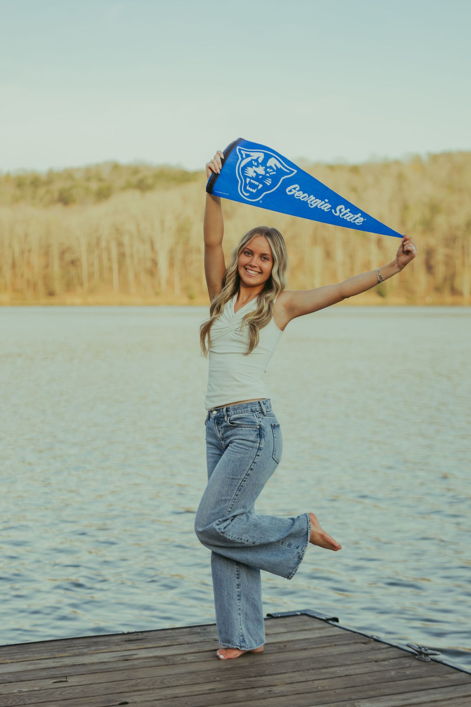 Smiling blonde girl holds Georgia State pennant flag on a dock over a calm lake.