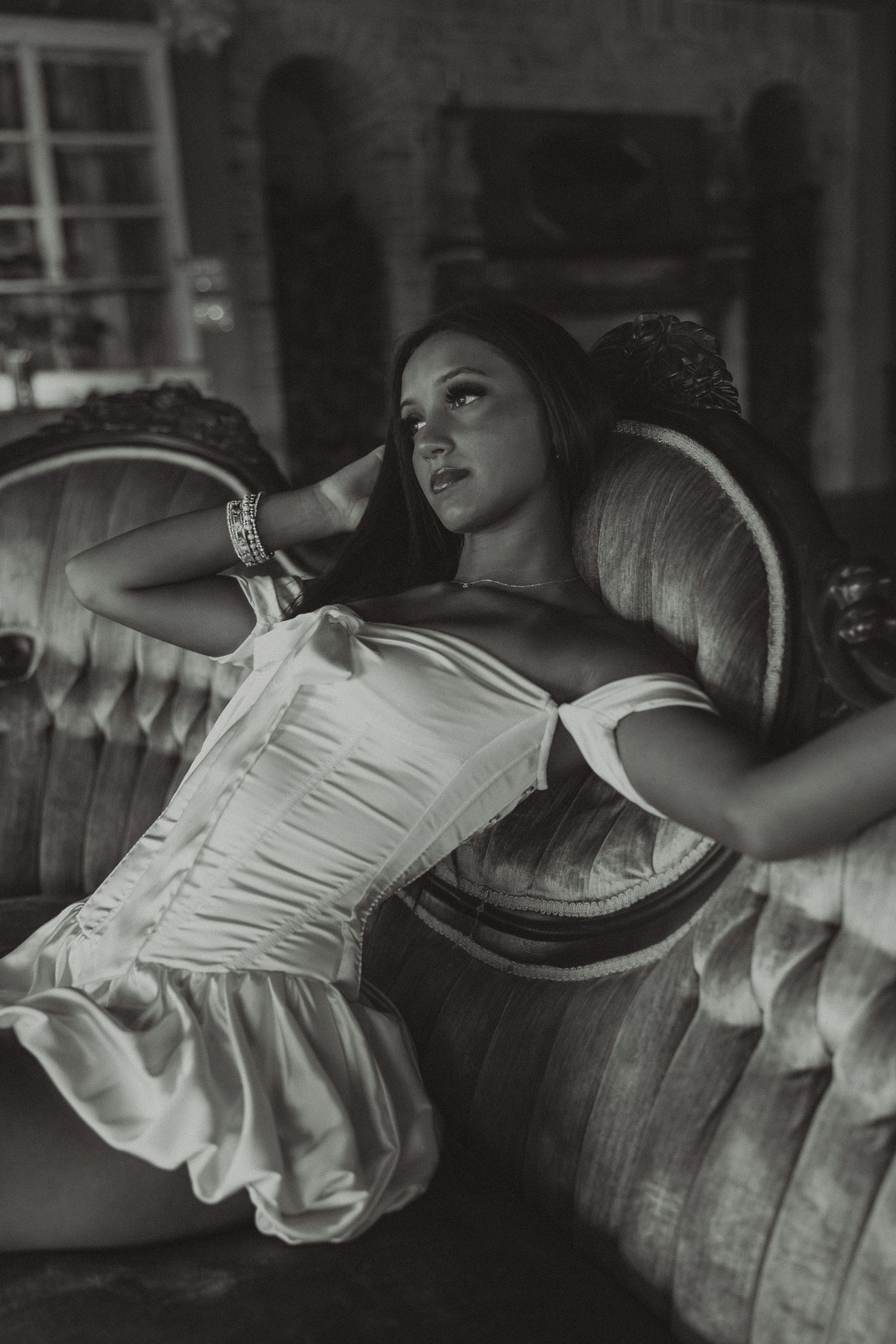 Black and white portrait of woman in white corset reclining on ornate vintage sofa.