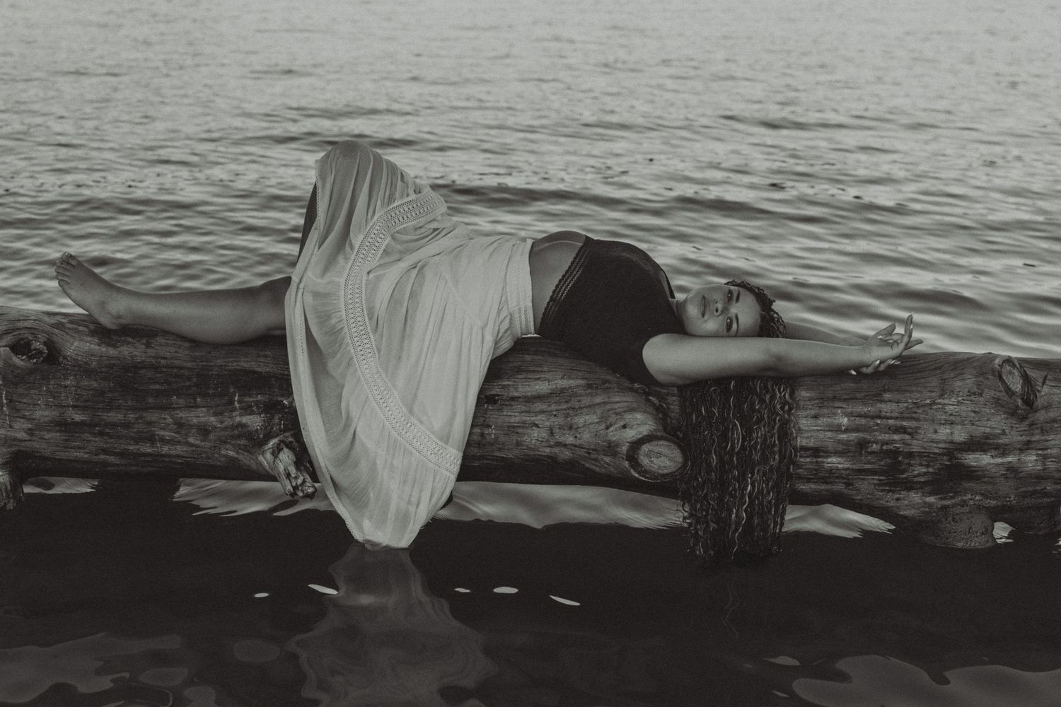 Black and white photo of woman in flowing dress lying on driftwood over water.