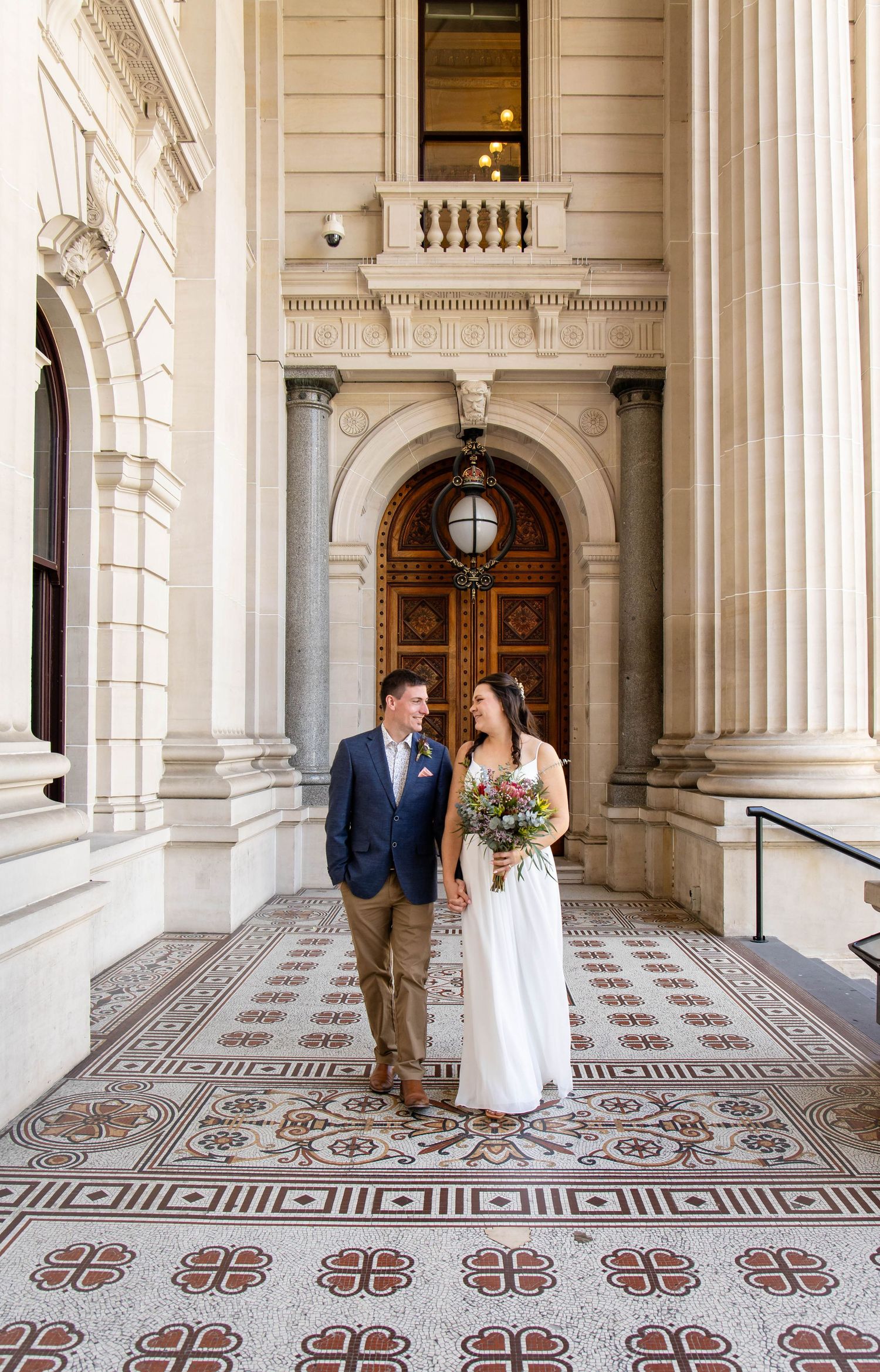 Couple walks through an elegant white stone hallway with ornate tiled floors featuring a repeating floral pattern.