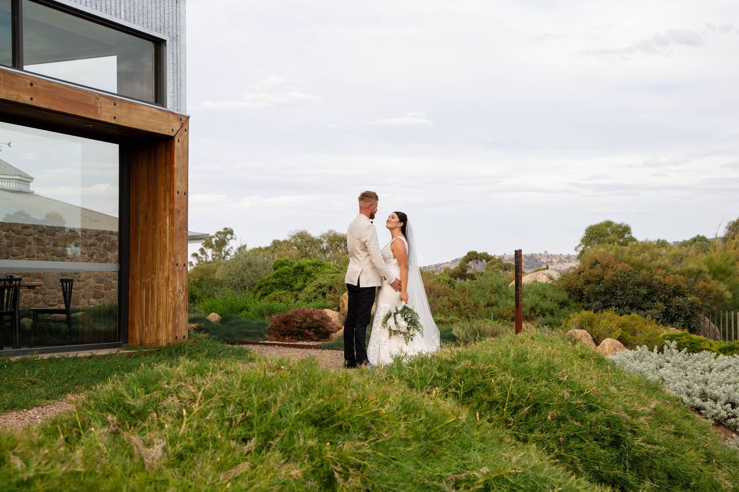 Wedding couple shares romantic kiss on rolling green hills overlooking scenic mountain landscape at sunset.