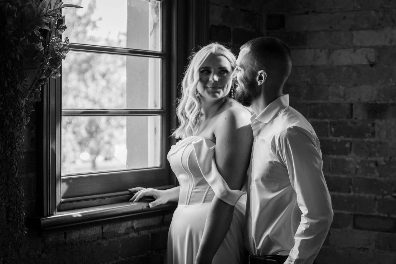 Romantic black and white photo of a couple in formal attire embracing by a window with natural light streaming in.