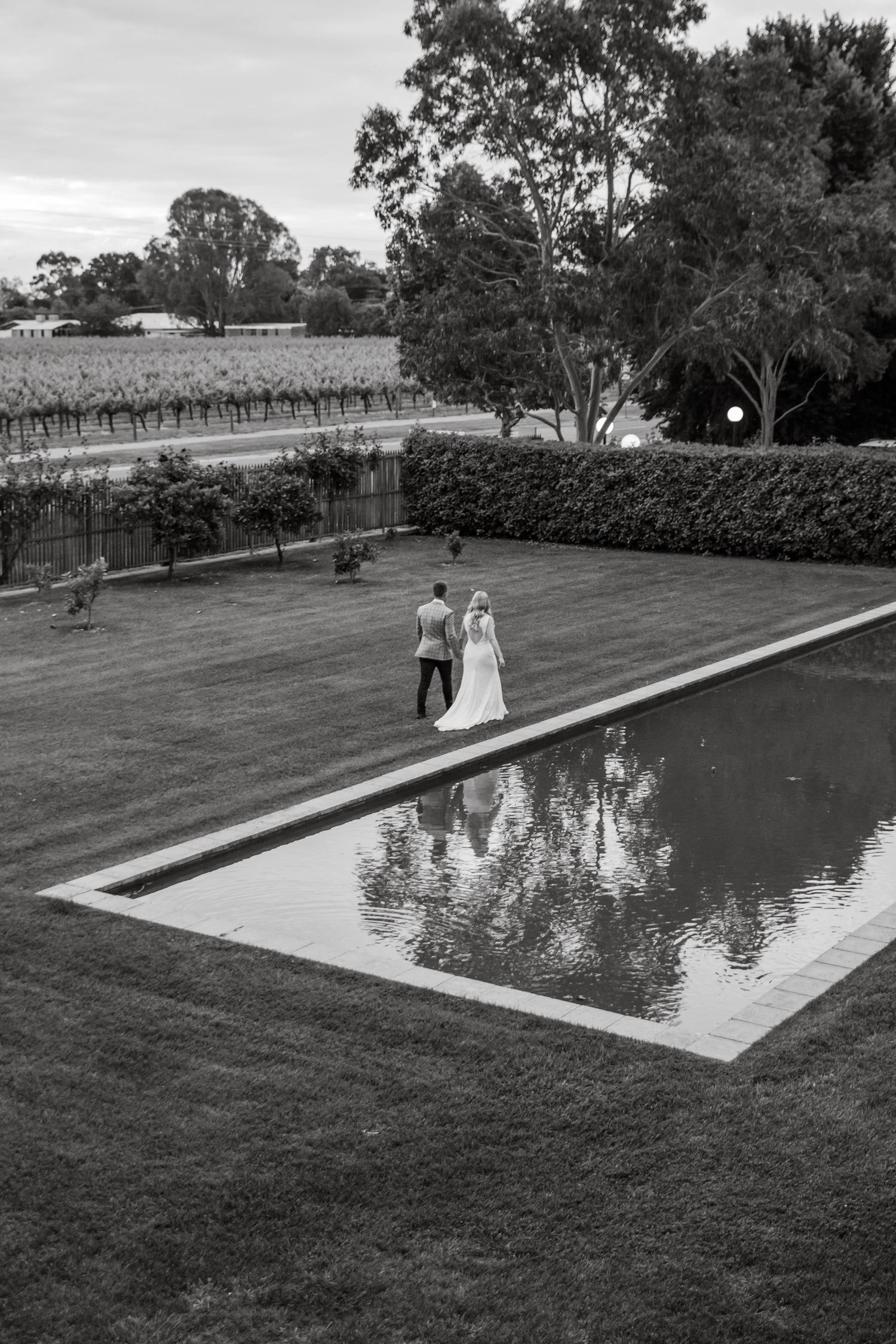 A black and white view of figures walking beside a reflecting pool at a vineyard estate with manicured gardens.