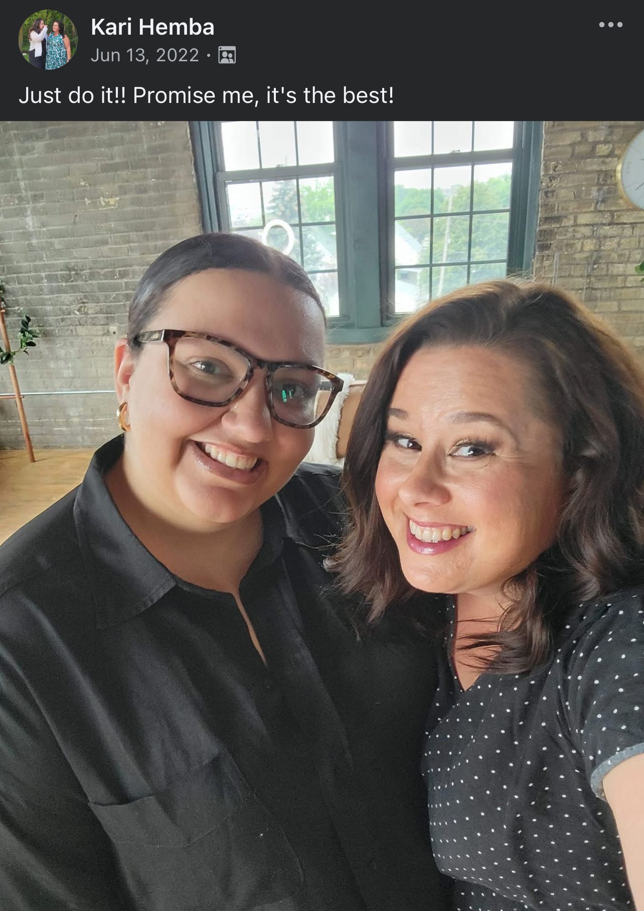 Two friends smile together in front of an industrial brick wall with large windows in a casual selfie.