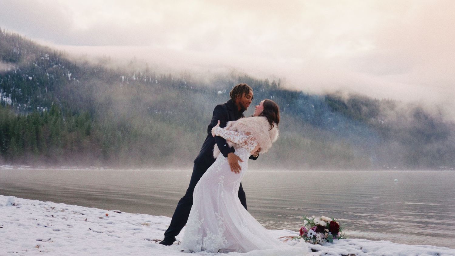 A romantic couple in wedding attire dances and embraces on a misty lakeside beach at sunset.
