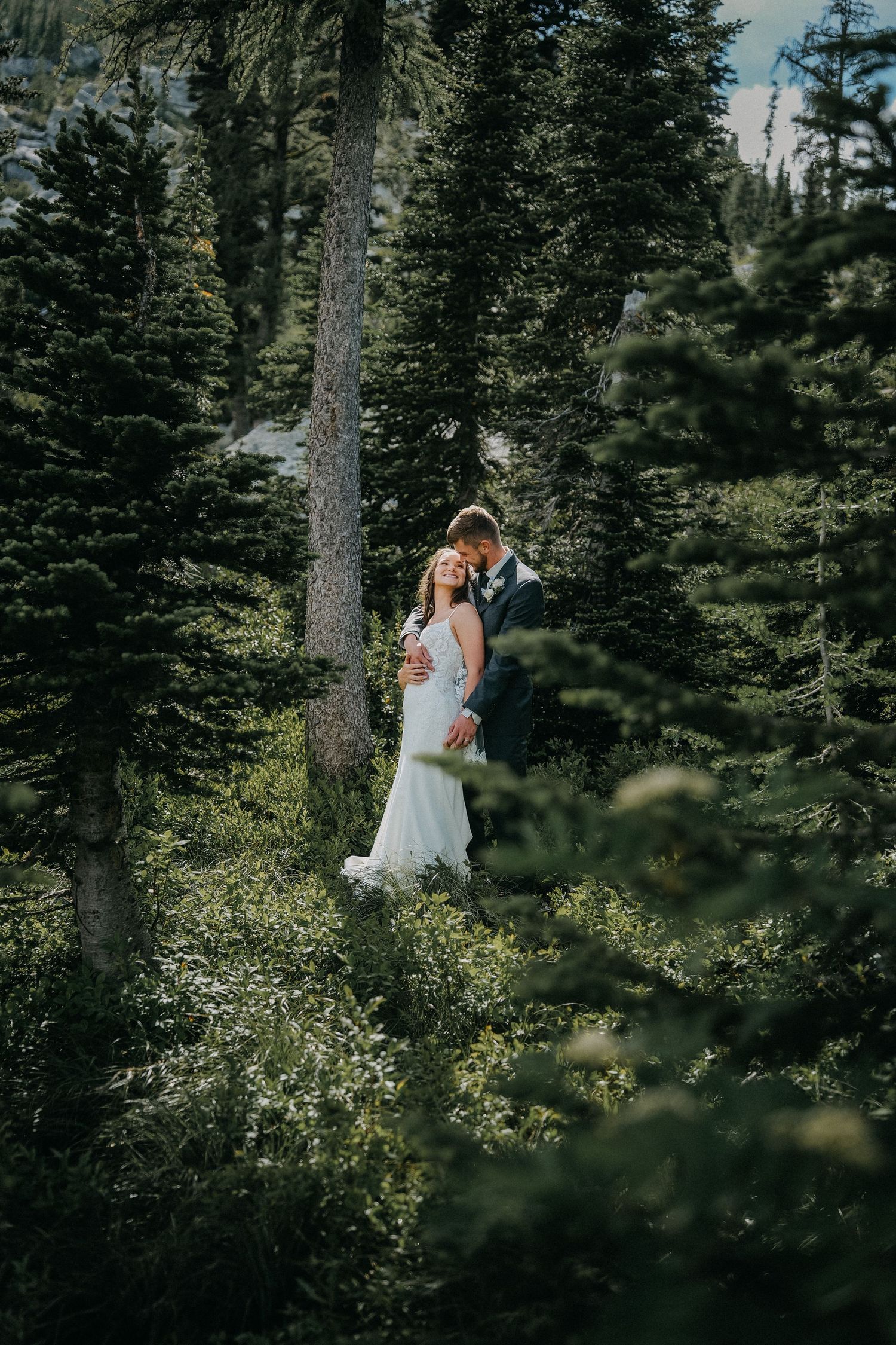 A couple in wedding attire shares an intimate moment among evergreen trees and forest foliage.