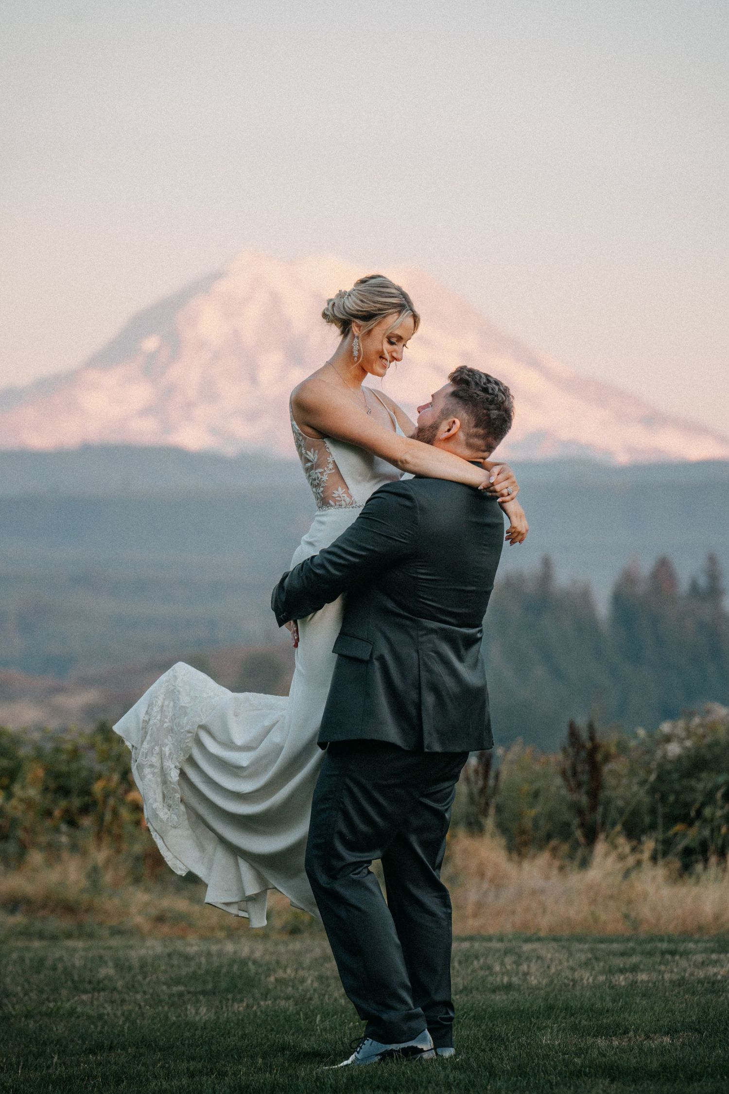 A couple in wedding attire embraces in front of a scenic mountain landscape at sunset.