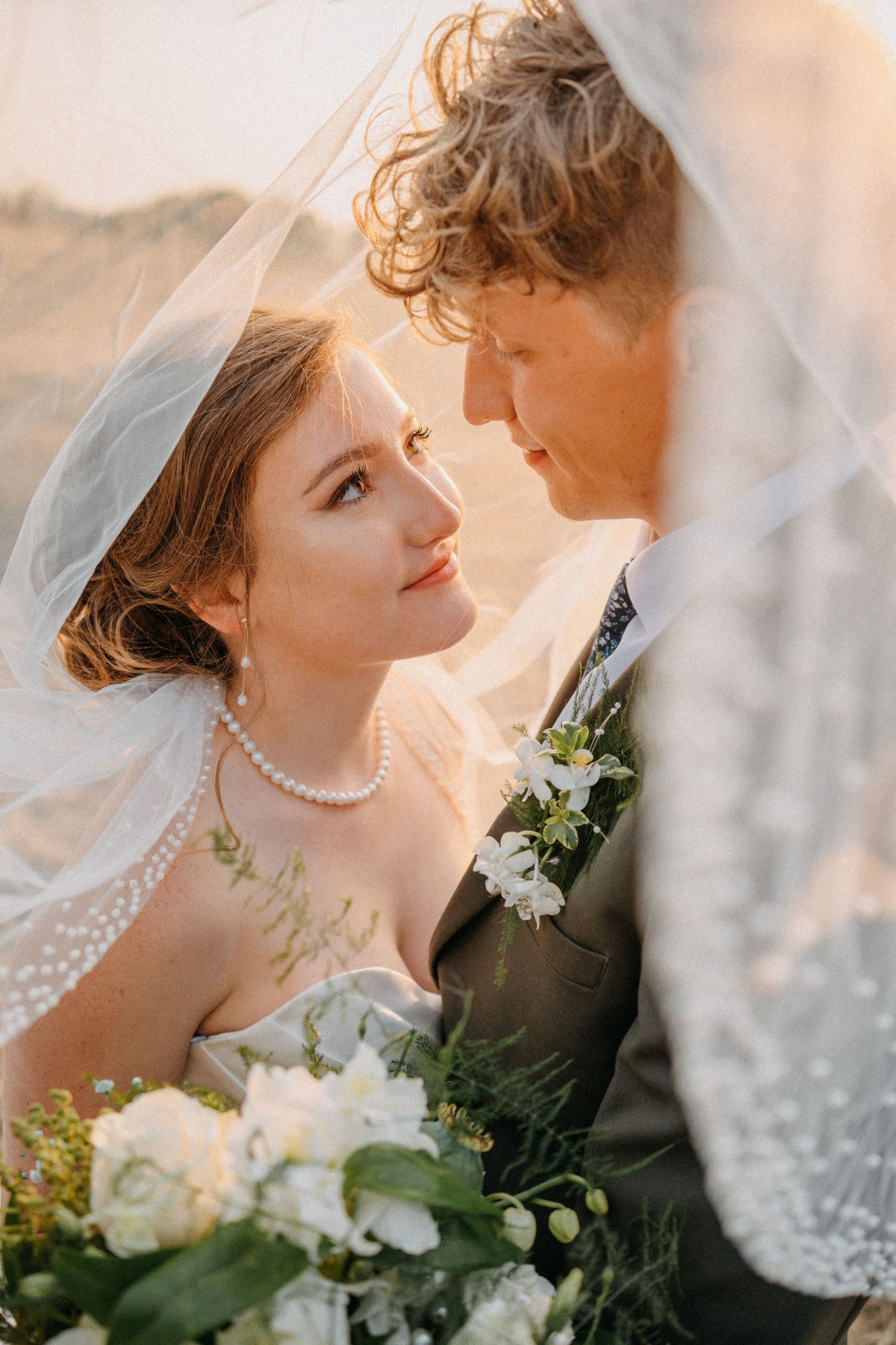 A romantic wedding portrait of a couple sharing an intimate moment under a flowing veil against warm sunlight.