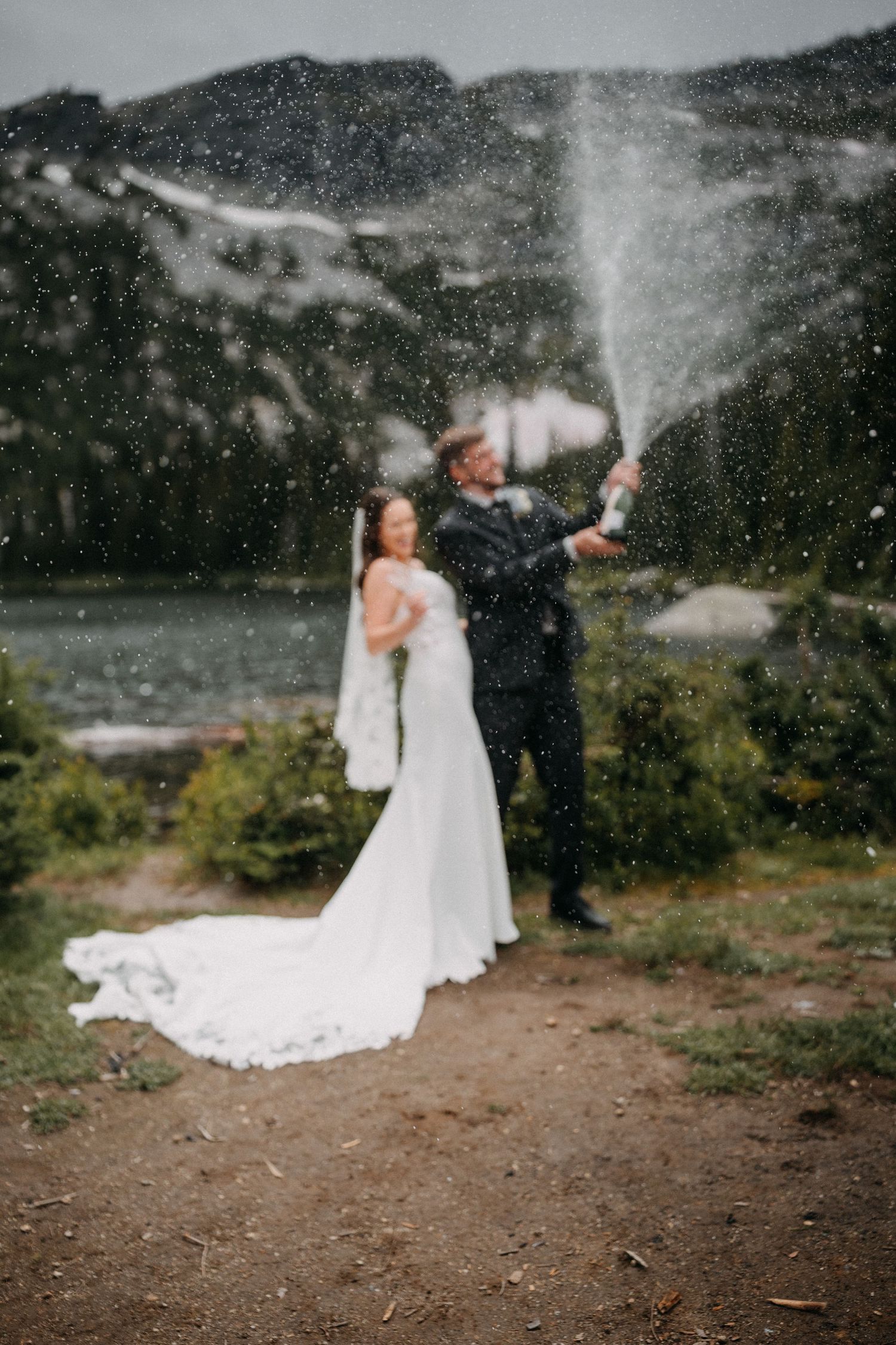 A couple celebrates with champagne sprays in a scenic mountain setting during their wedding photoshoot.