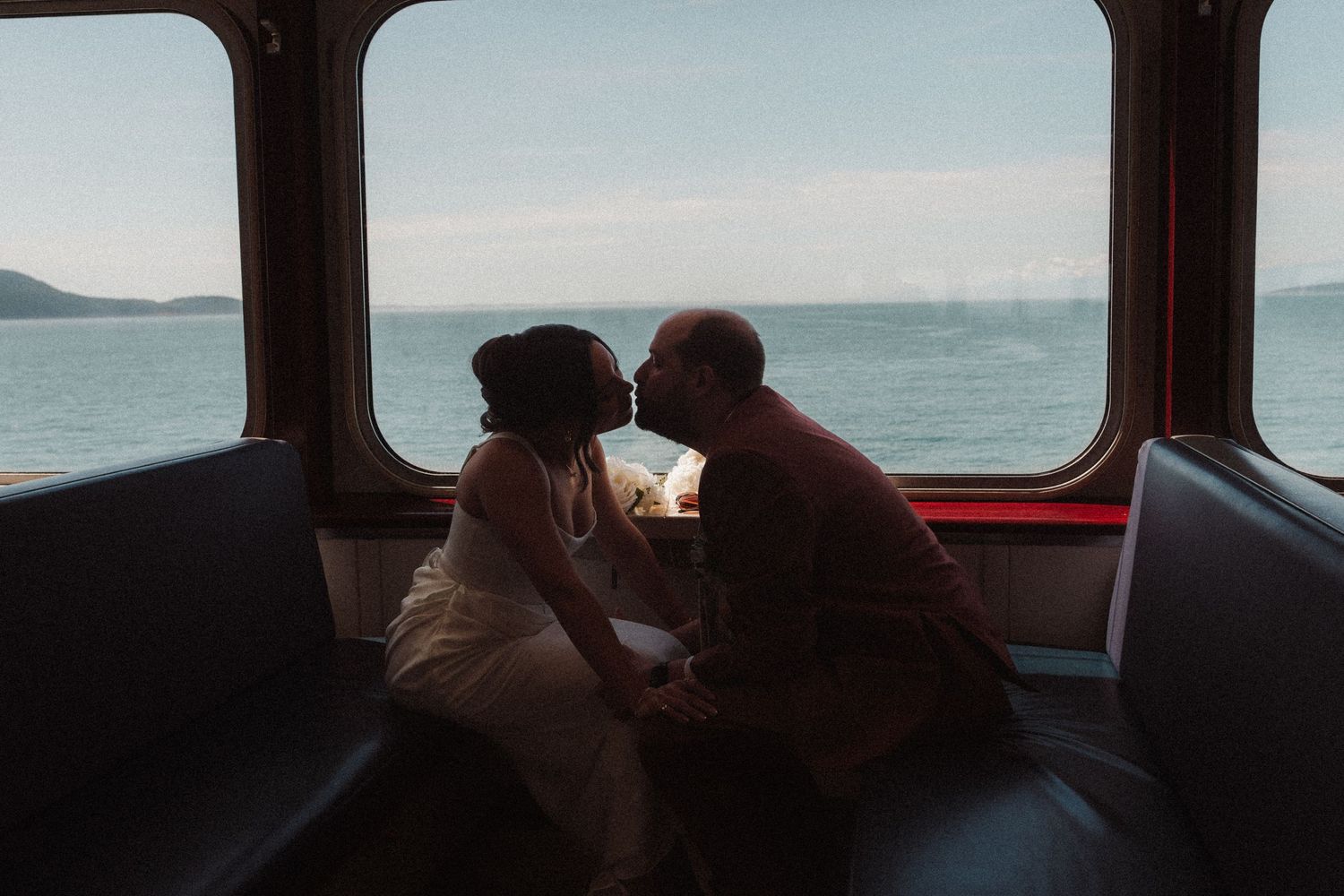 Silhouetted couple in wedding attire share a kiss on a ferry bench, with ocean views and distant hills through the large windows.