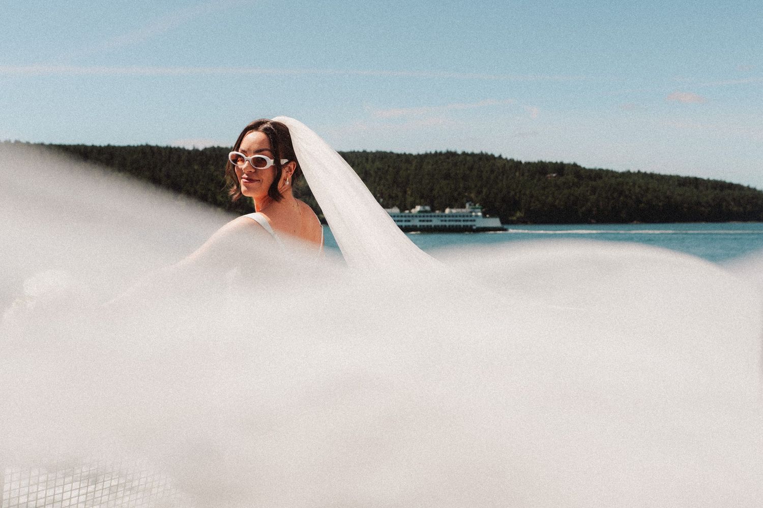 Bride in white sunglasses and a flowing veil billowing in the wind, standing near the water with a ferry and forested island behind her.