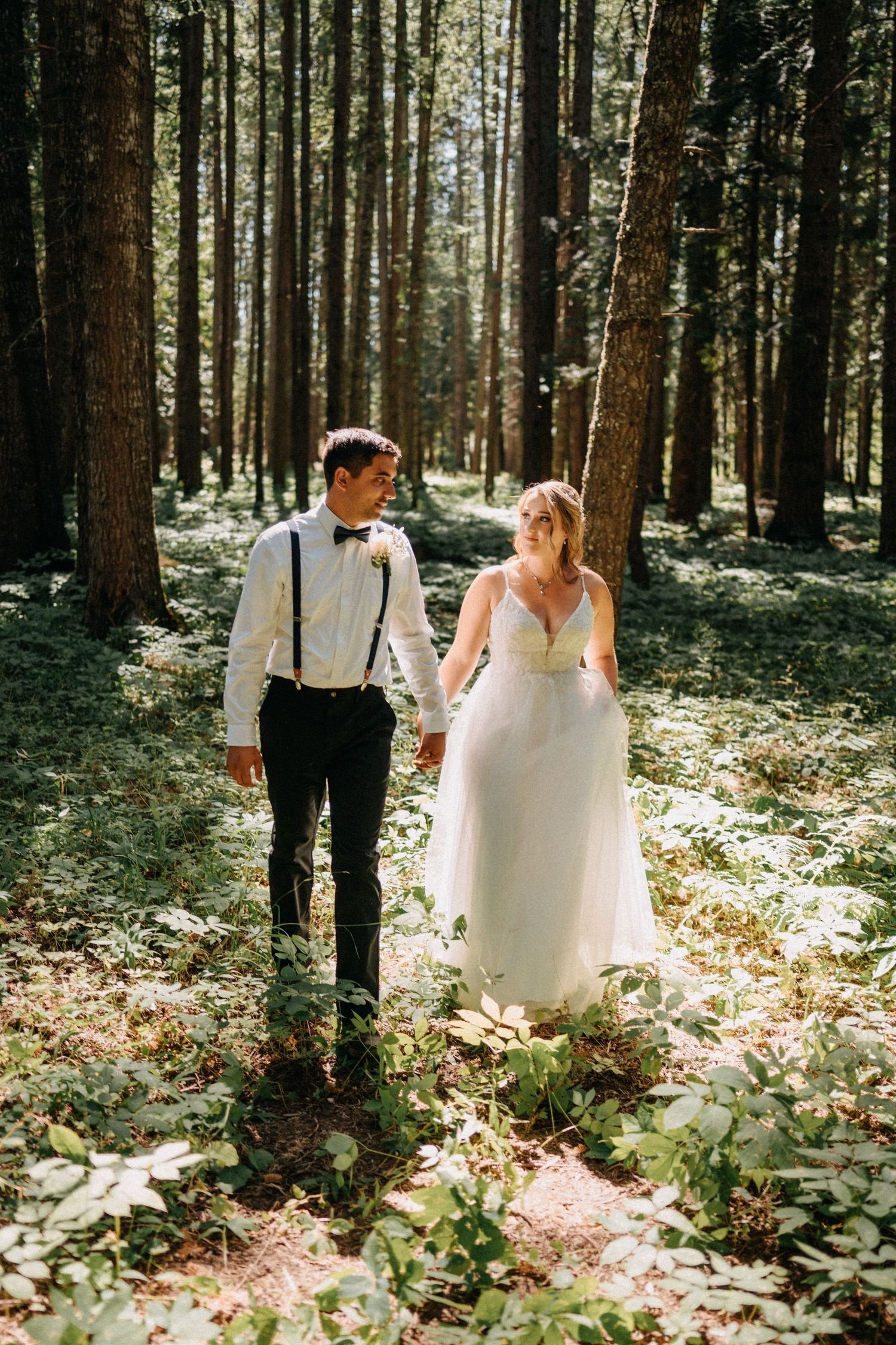 A romantic wedding couple walks through a sunlit forest with tall trees and scattered leaves on the ground.