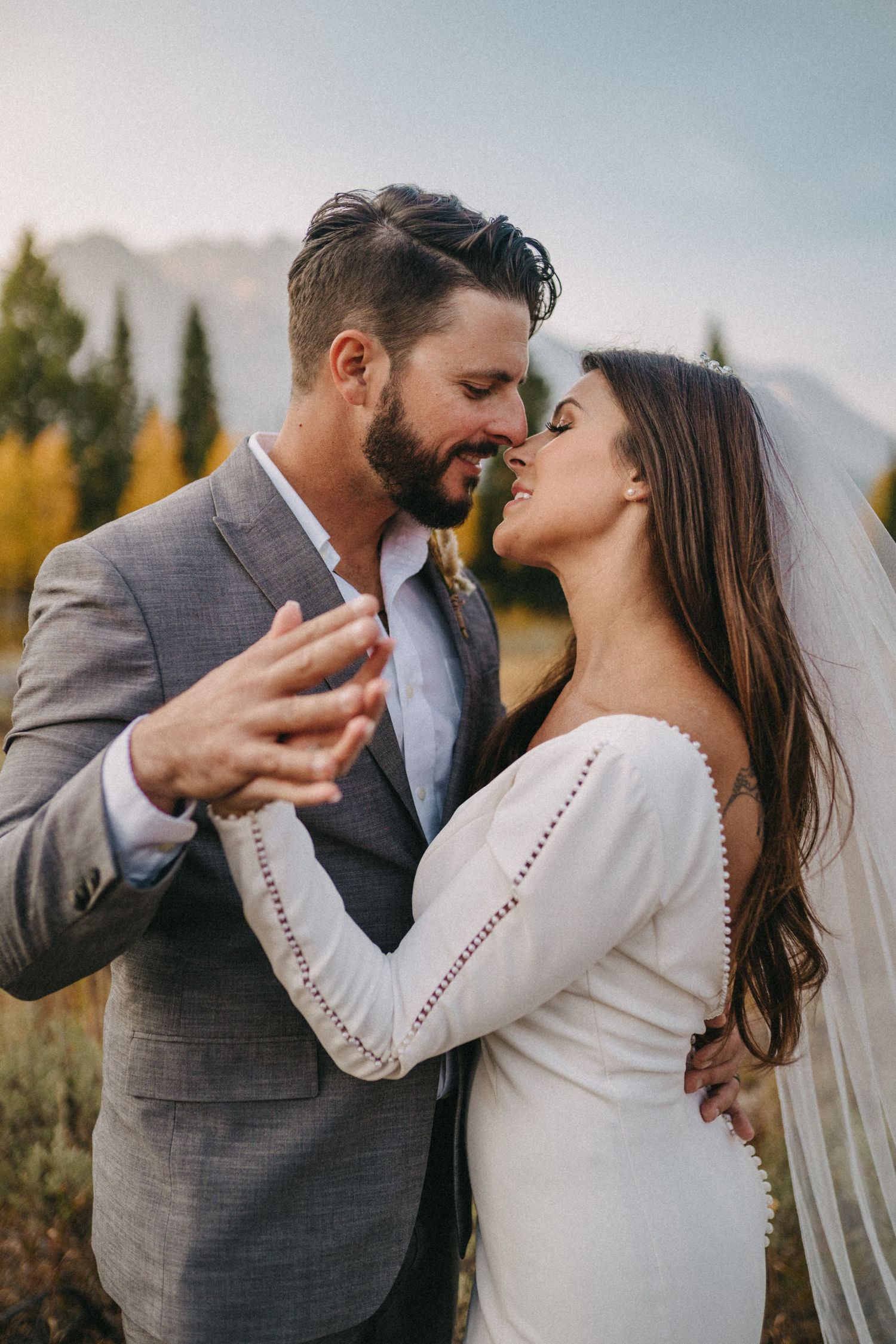 Romantic wedding couple sharing intimate kisses in outdoor autumn setting wearing grey suit and white dress.