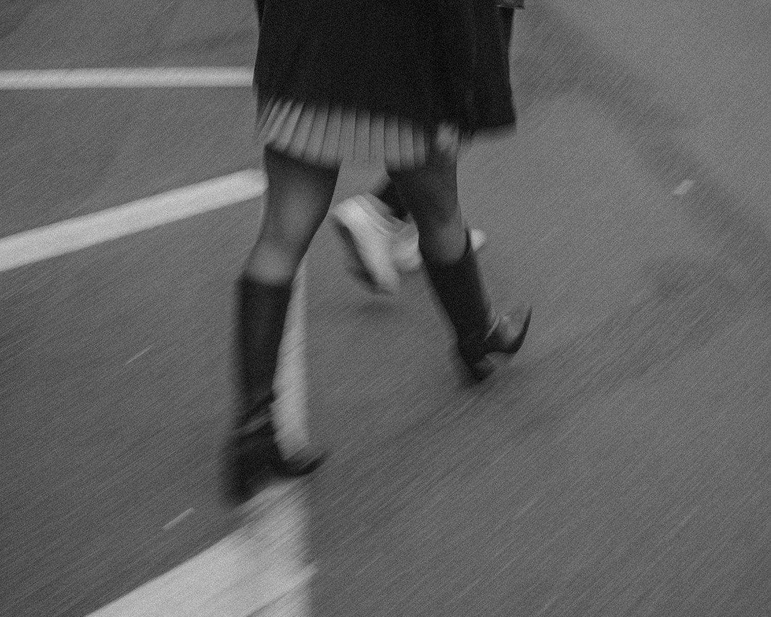Blurred black and white photo of a person in a skirt and knee-high boots walking across a wet street.