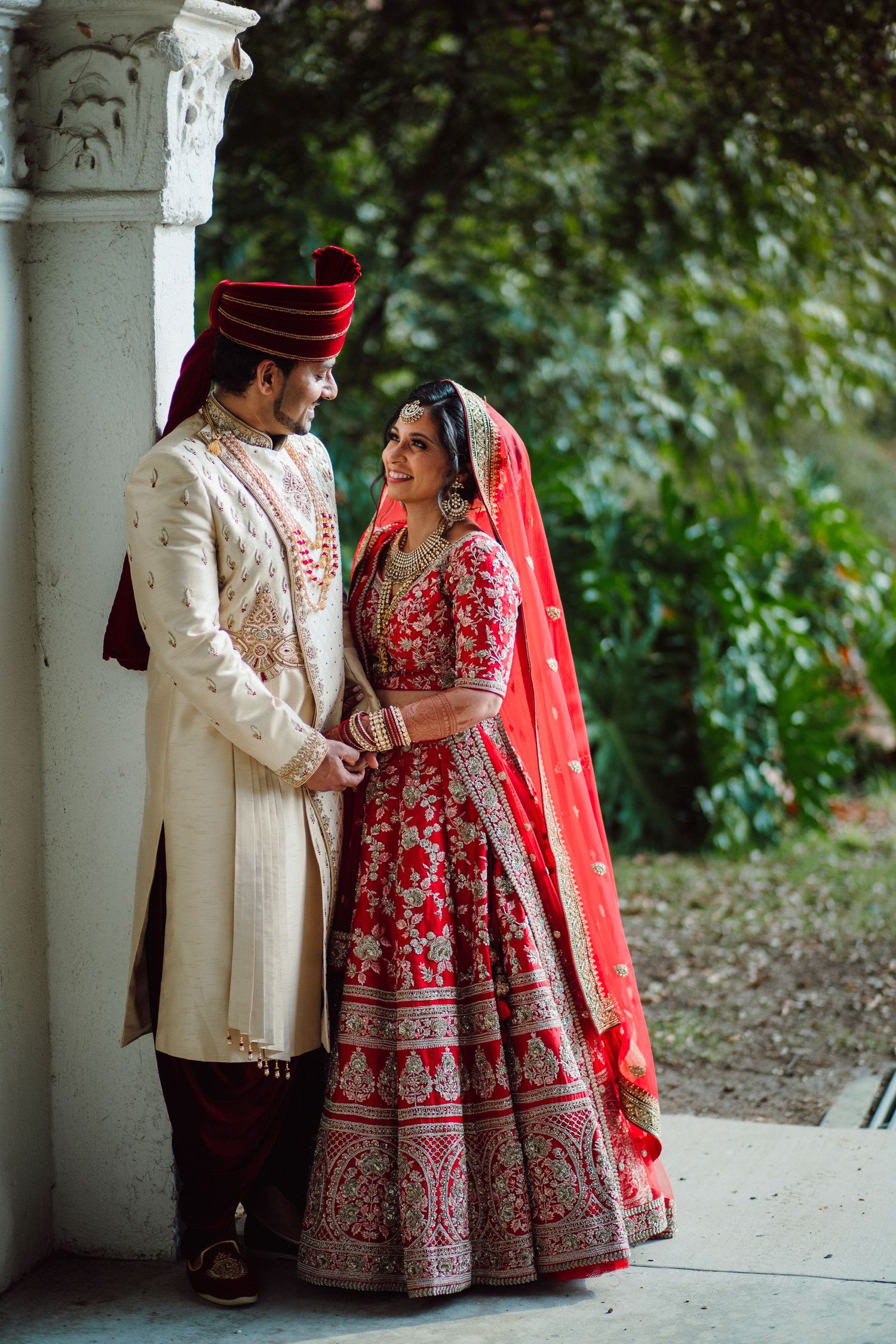 Indian bride in red lehenga and groom in cream sherwani share a romantic moment at their traditional wedding.