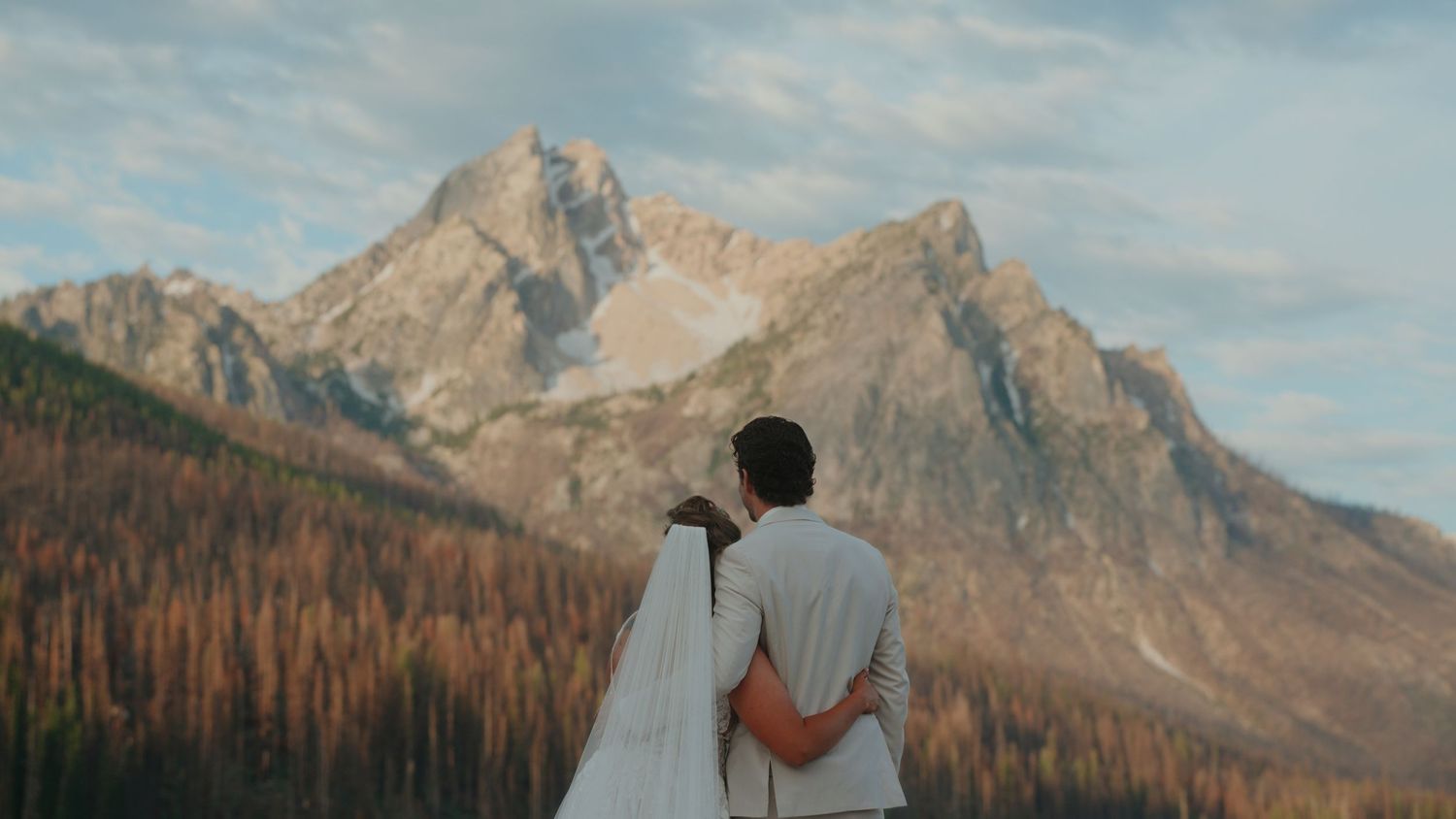 Newlywed couple embracing in front of a rugged mountain peak with an autumn forest below, the bride's white veil flowing behind her.