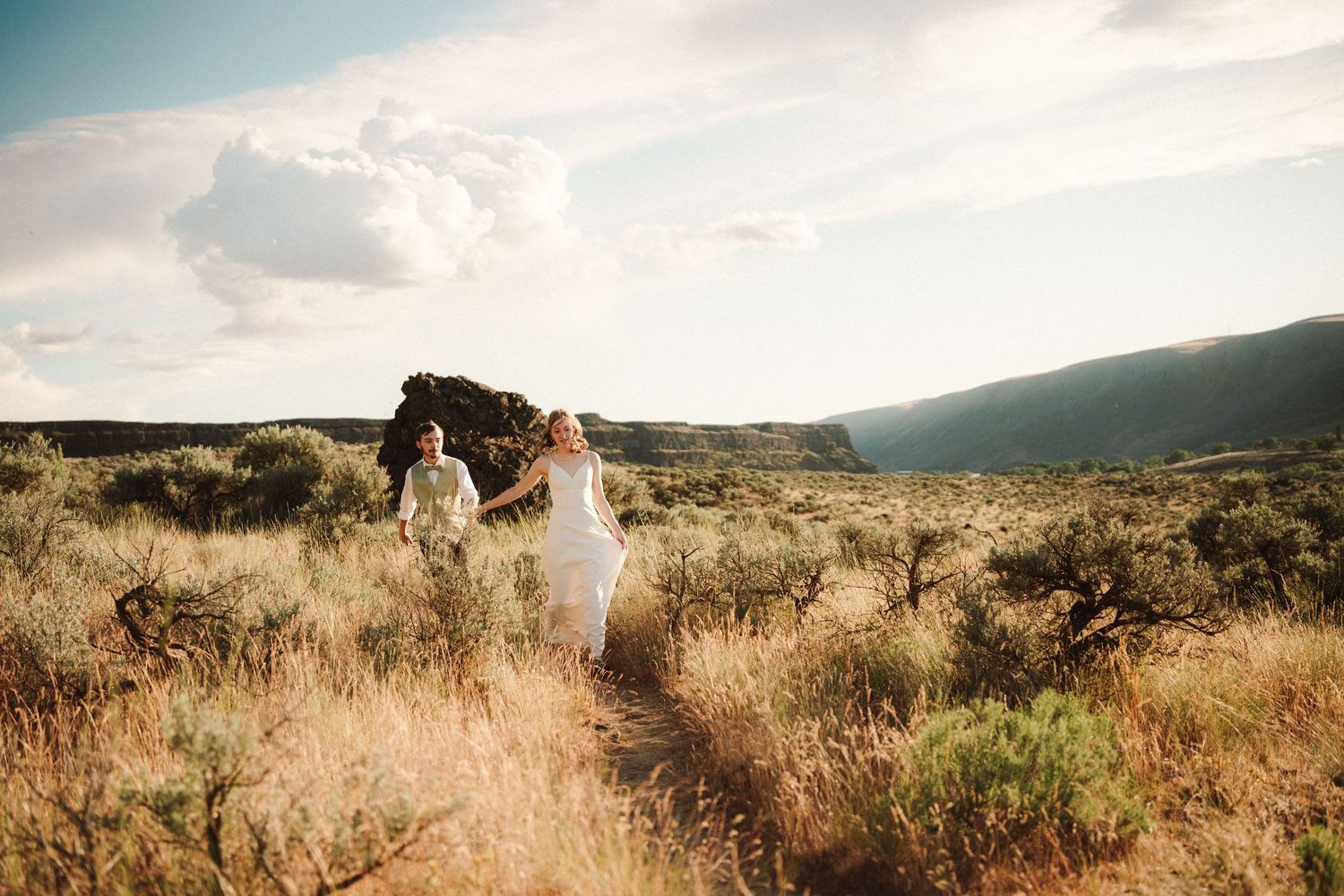 Bride in a white gown and groom hold hands while walking through golden sagebrush grassland with rolling hills and a bright cloudy sky.