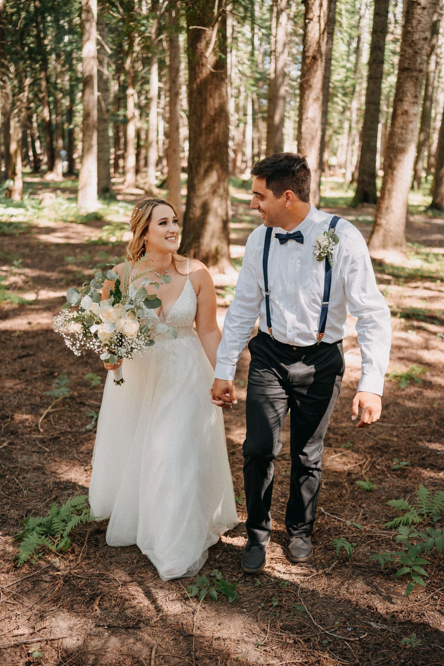 A couple walks hand in hand through a sunlit forest wearing wedding attire.