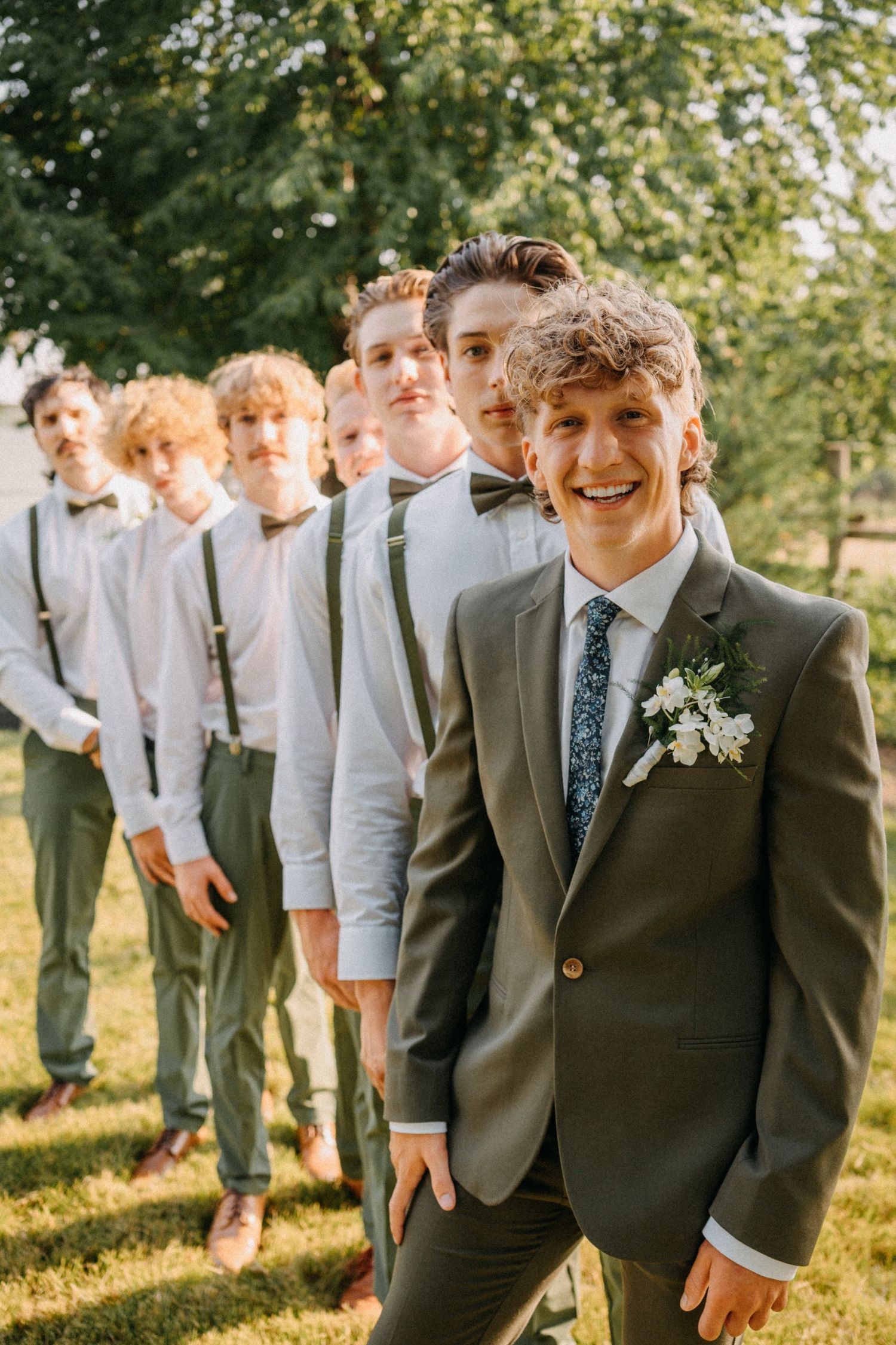 Smiling groom in gray suit with boutonniere stands before groomsmen in white shirts and suspenders outdoors.