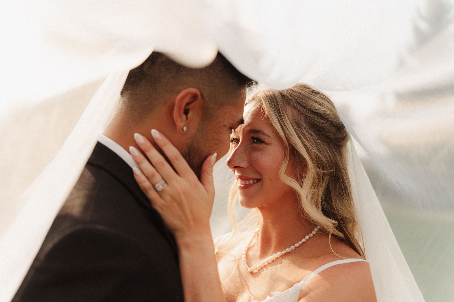 Smiling bride in a pearl necklace and groom in a dark suit share an intimate moment beneath a sheer white veil on their wedding day.