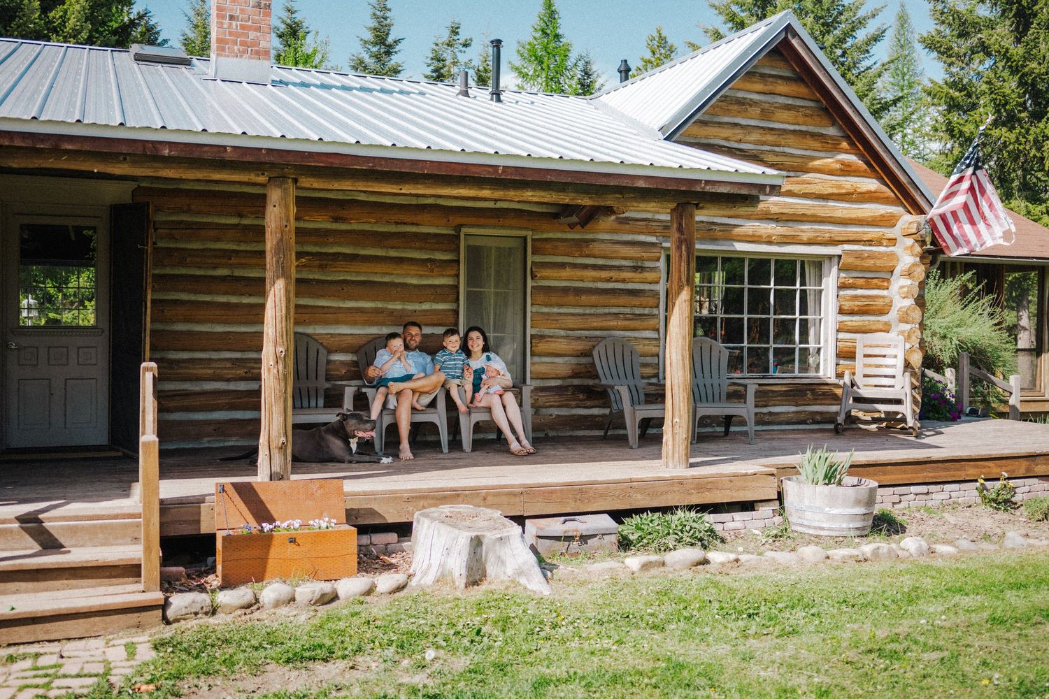 Family relaxing on the porch of a rustic log cabin with metal roof, surrounded by trees and an American flag.