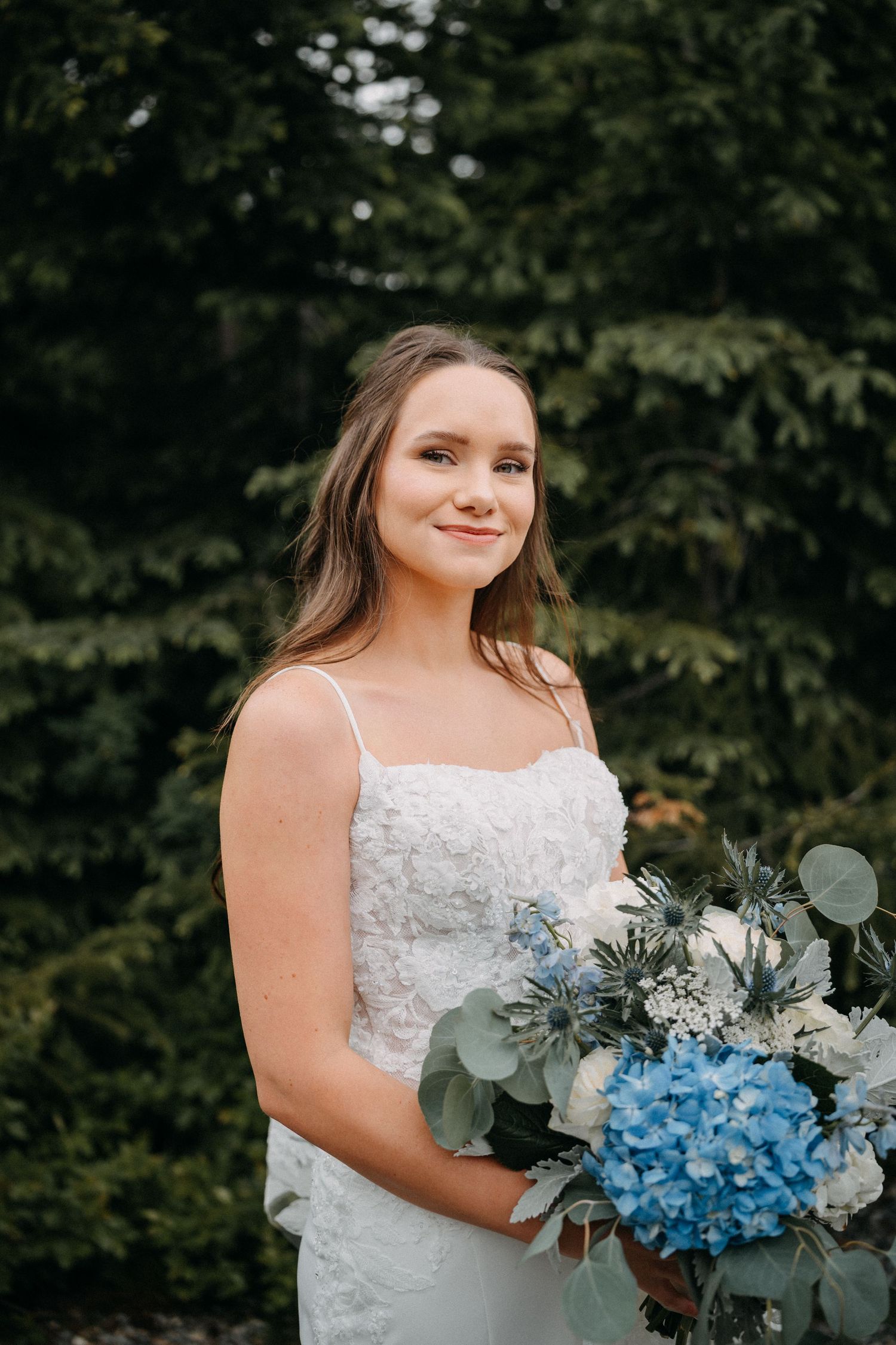 A bride in a white lace dress holds a blue hydrangea and eucalyptus bouquet against a dark forest backdrop.