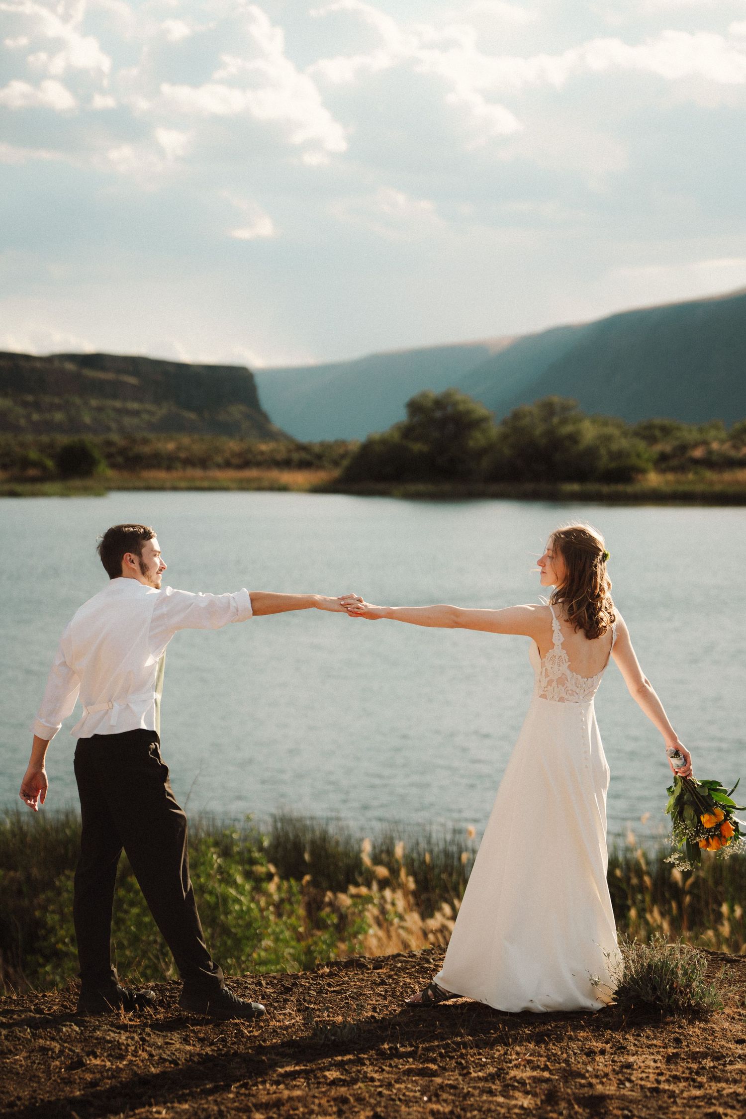 Bride in a white gown holding a bouquet and groom in a white shirt hold hands by a calm lake with hills in the background.
