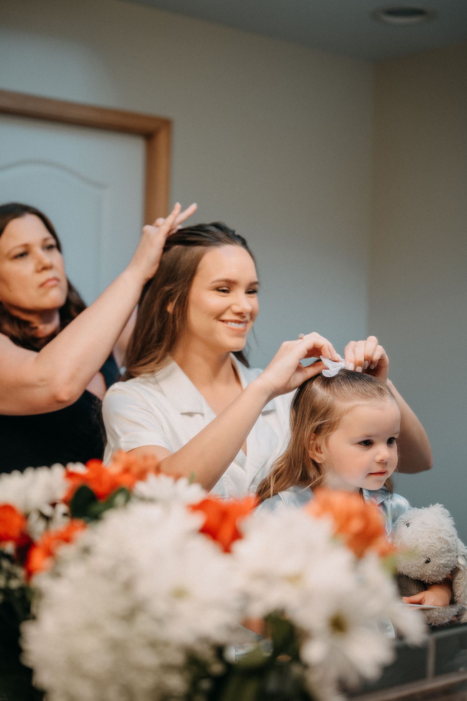Wedding preparations scene with white and orange flowers in foreground while stylists work on hair in the background.