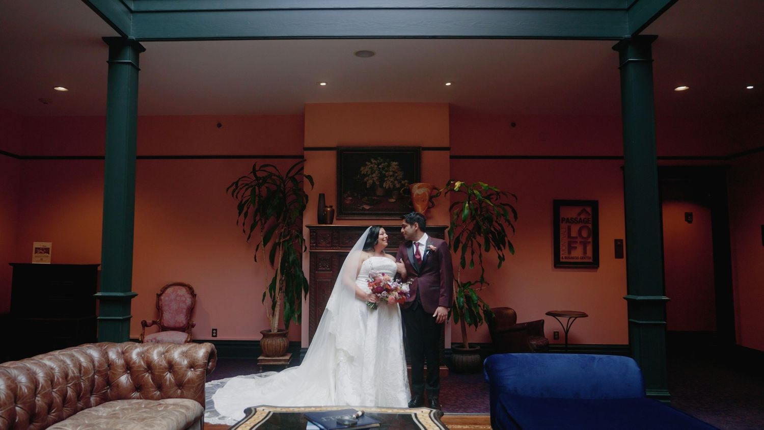 Bride and groom share a romantic moment in an elegant coral-walled venue with lush greenery and vintage blue velvet sofa.