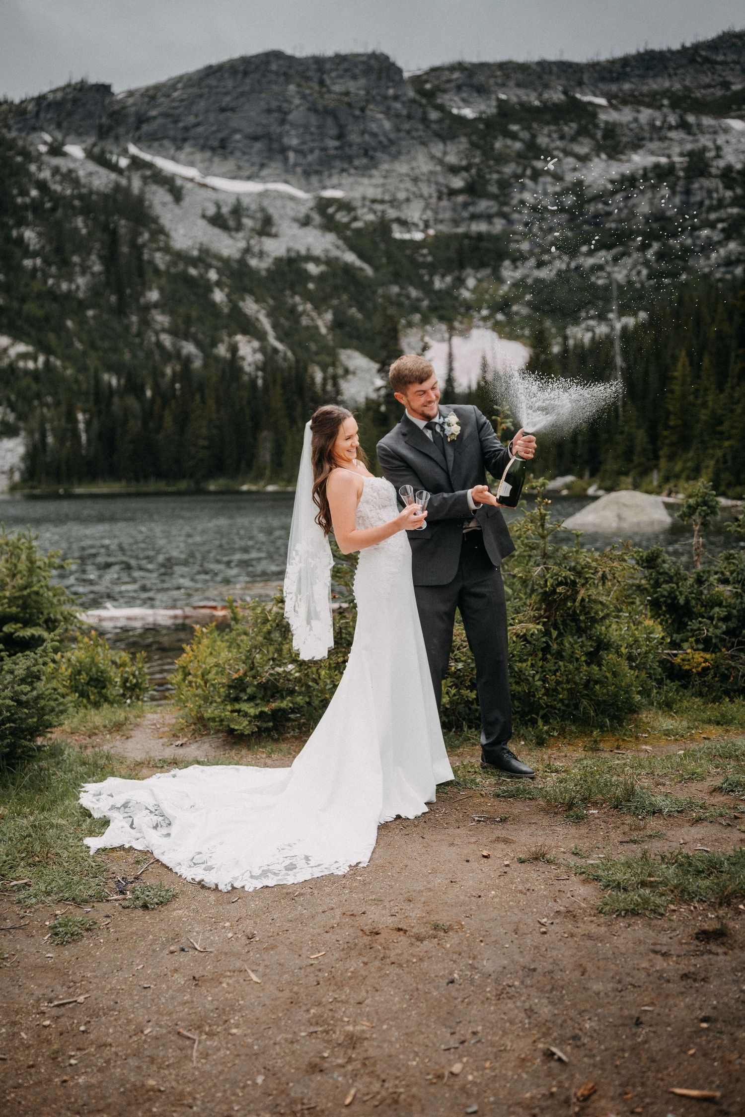 Bride in a long lace gown and veil watches groom pop champagne beside a mountain lake with rocky peaks and evergreen forest.
