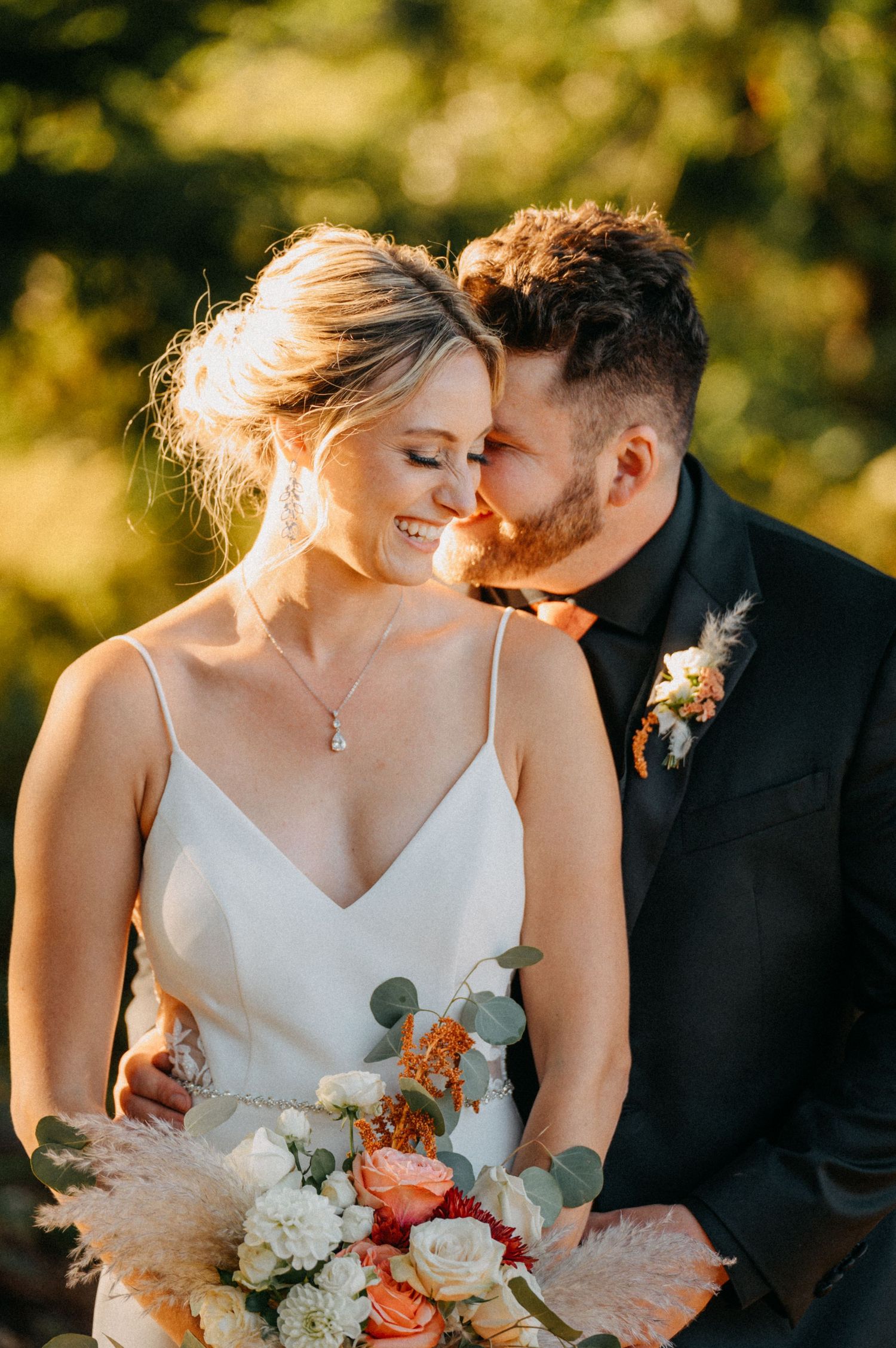 A romantic wedding couple shares intimate moments in golden sunlight while holding a bouquet of white and peach flowers.