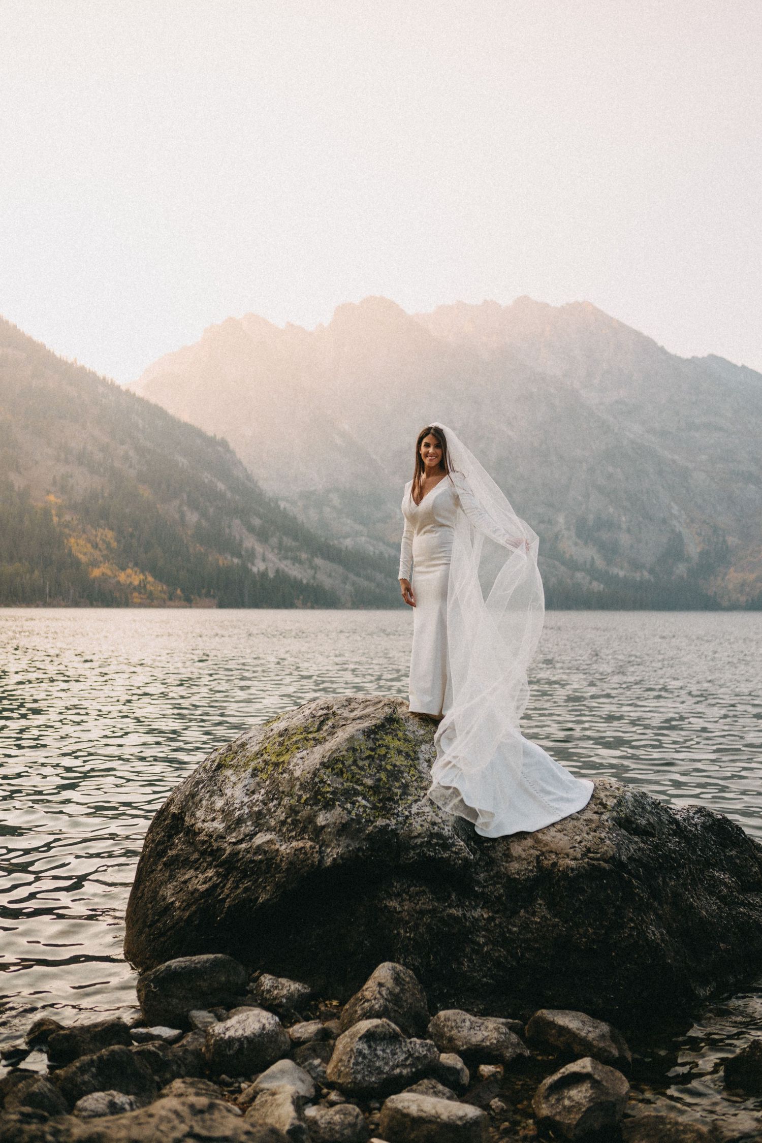 A person in a flowing white dress stands on a rock by a mountain lake at sunset, with dramatic peaks in the background.