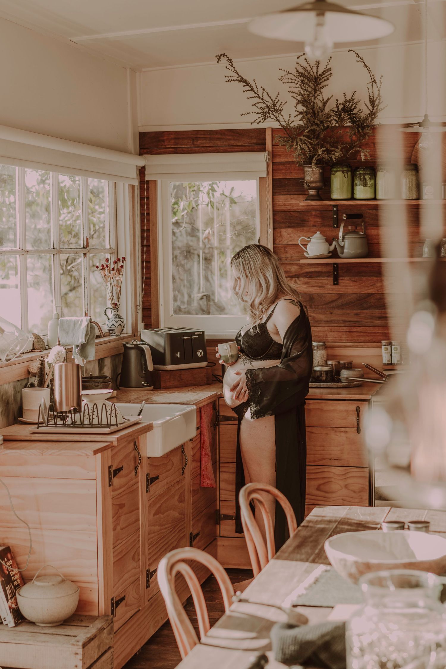 Blonde woman in black robe standing in rustic wooden kitchen holding a cup near window.