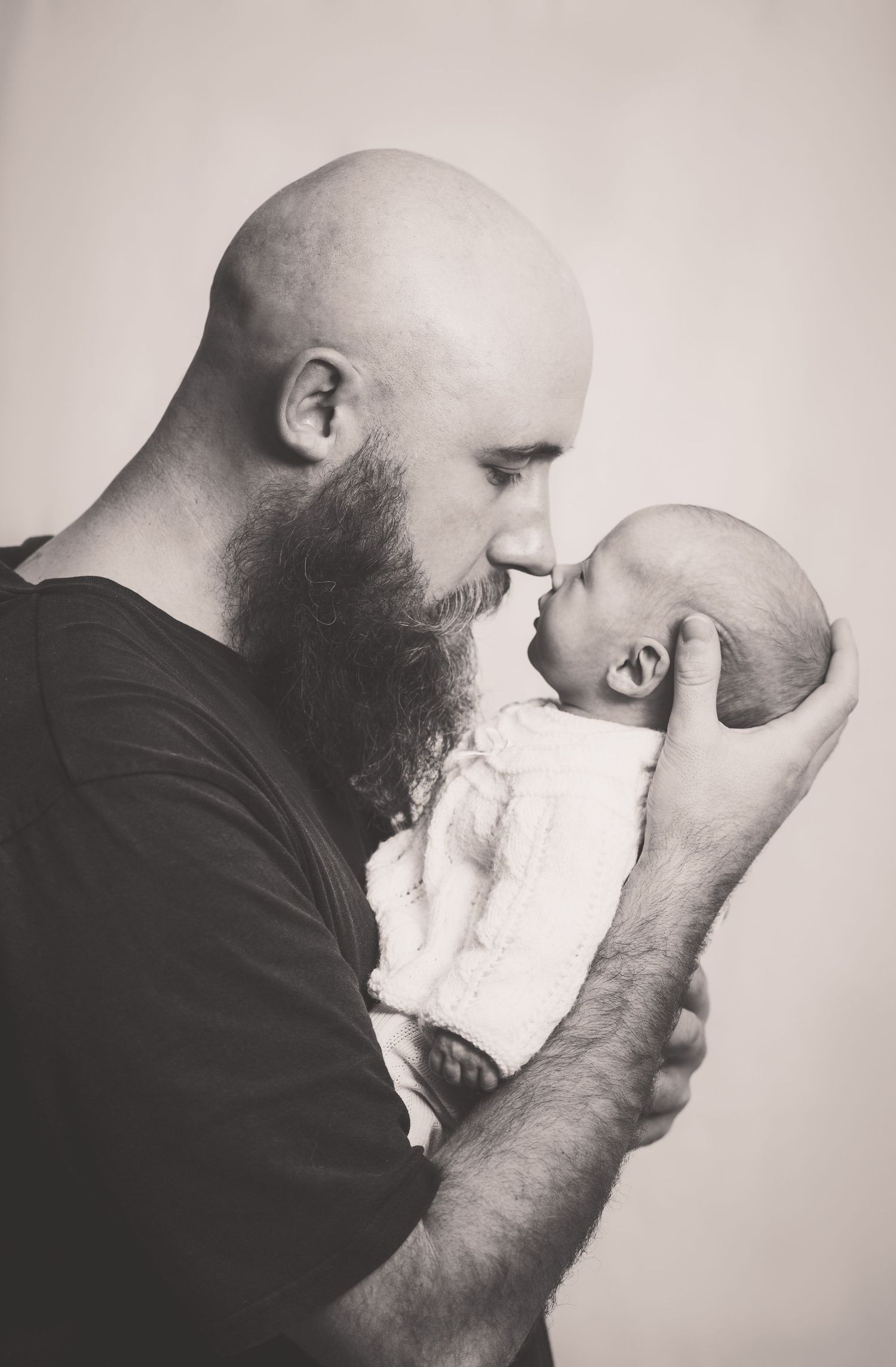 Bald bearded father holding newborn baby face to face in black and white portrait.